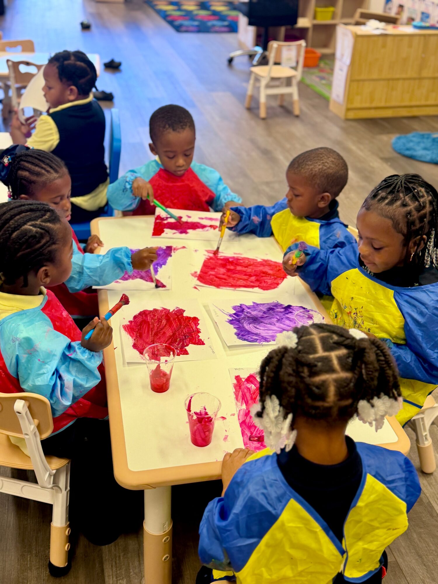 Children sit around a table painting with colorful paints in a classroom, with chairs and educational materials visible in the background.