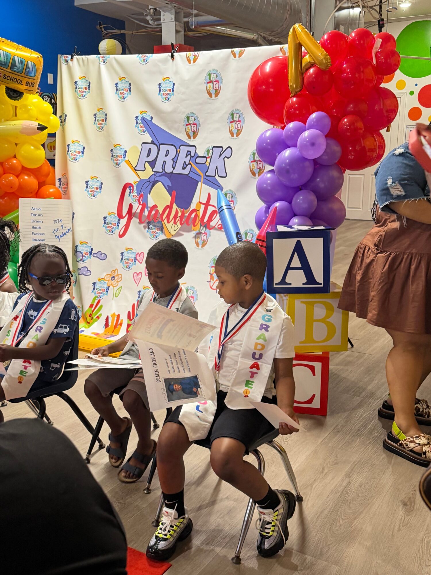 Two children sitting on chairs reading a newspaper at a colorful event with balloons and a decorated backdrop.