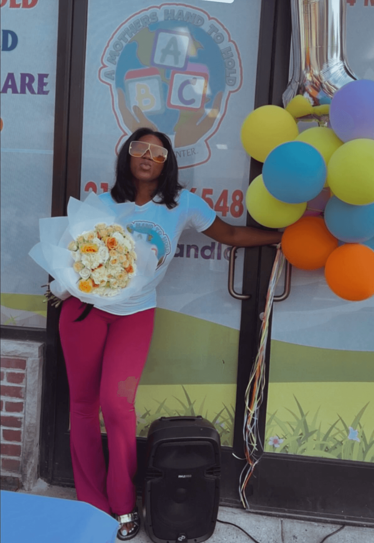 Woman holding a bouquet of flowers, standing outside a building with colorful balloons and a logo in the background.