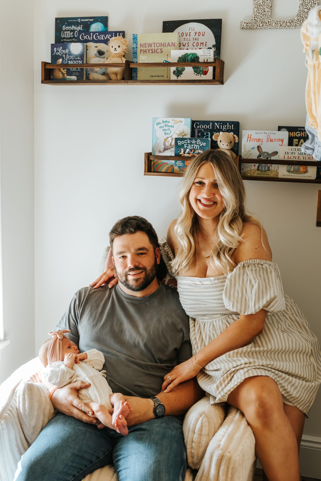 A smiling woman with blonde hair in a striped dress sits beside a man with a beard, holding a small dog, in a cozy room with bookshelves.