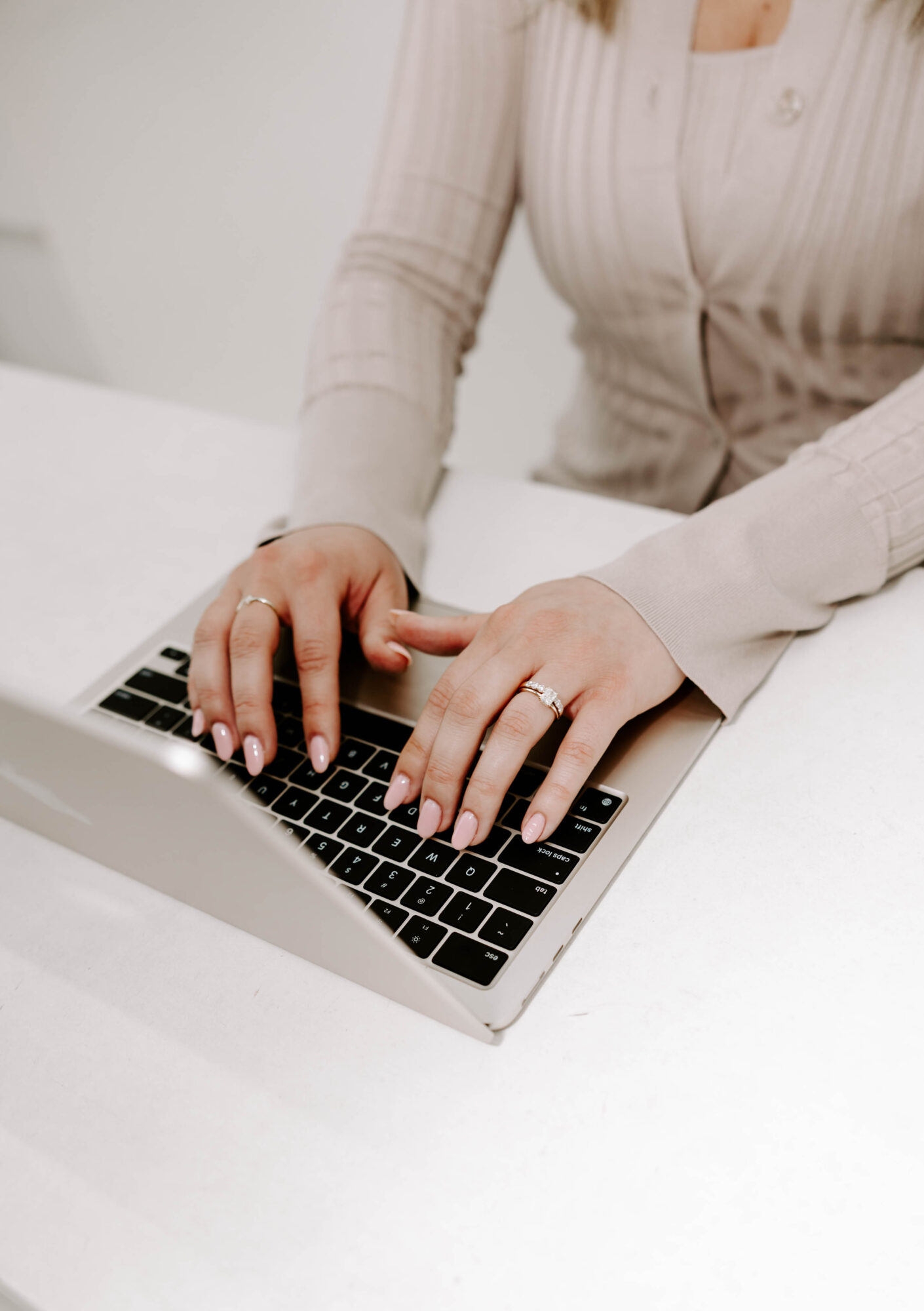 Person typing on a laptop with hands on keyboard, wearing a light-colored, long-sleeved shirt, sitting at a white surface.