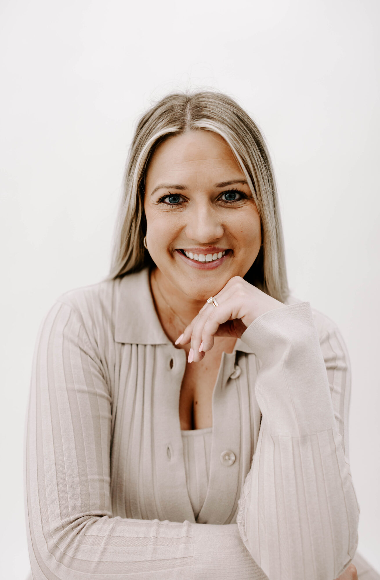 Young woman with long blonde hair smiling, resting her chin on her hand, wearing a light-colored shirt, against a plain white background.