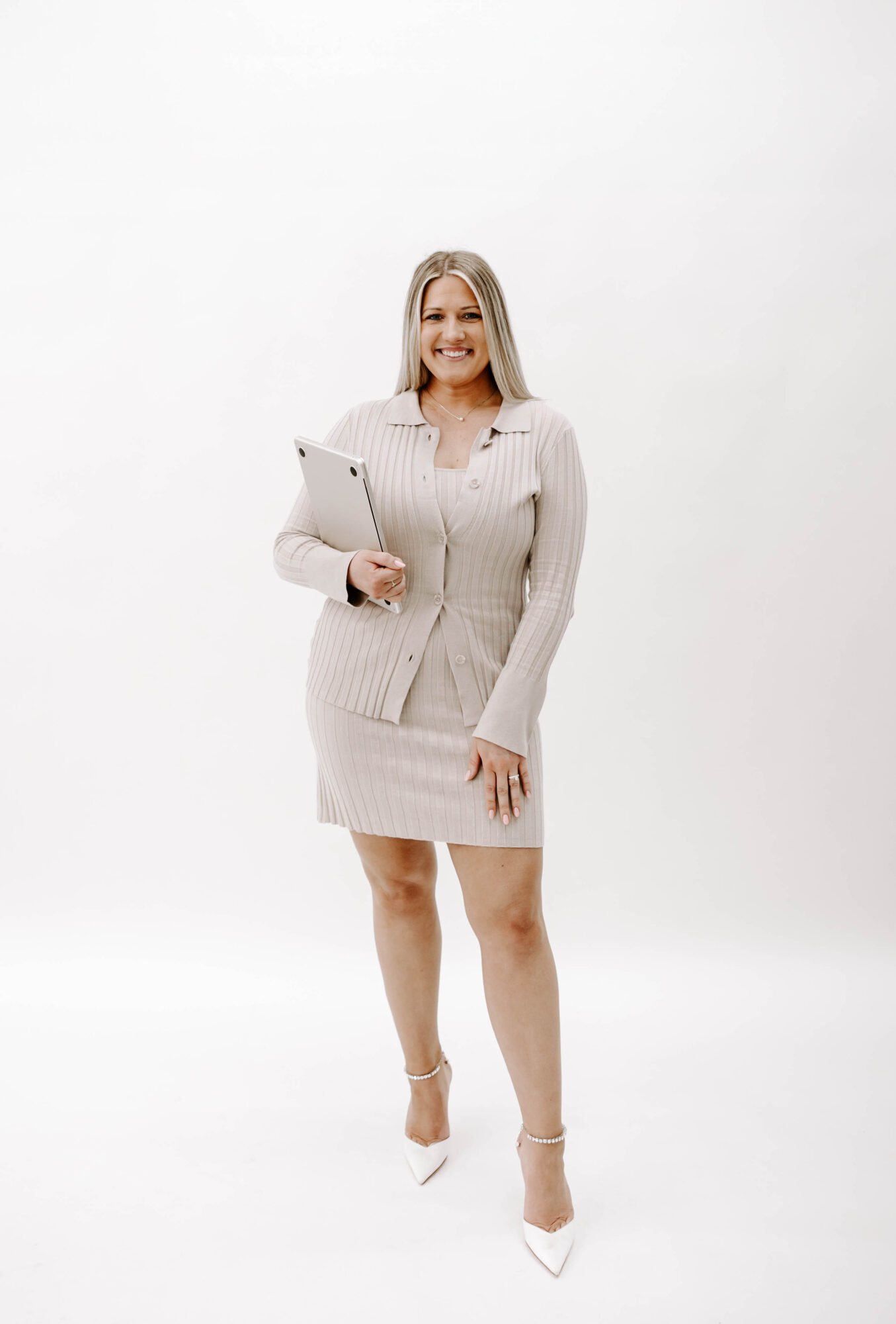 Woman in a beige dress holding a tablet, smiling, standing against a white background.