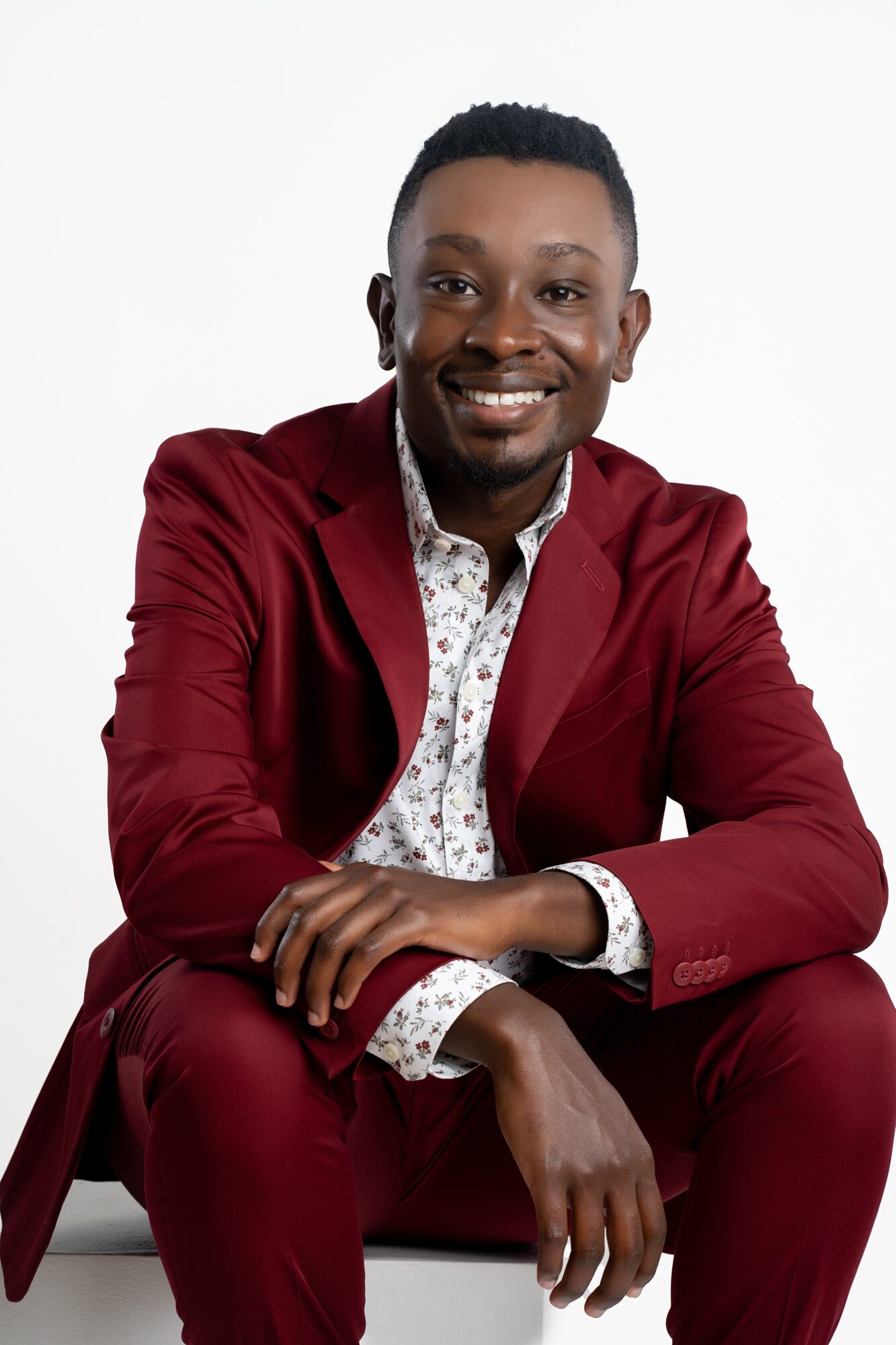 Smiling man in a red suit and patterned shirt sitting against a white background.
