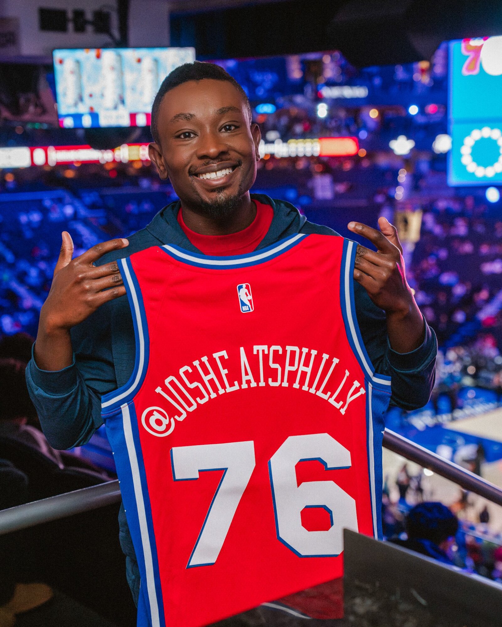 Person smiling and pointing at a red sports jersey with the number 76 and '@JUSHEATSPHILLY' on it, in a stadium.