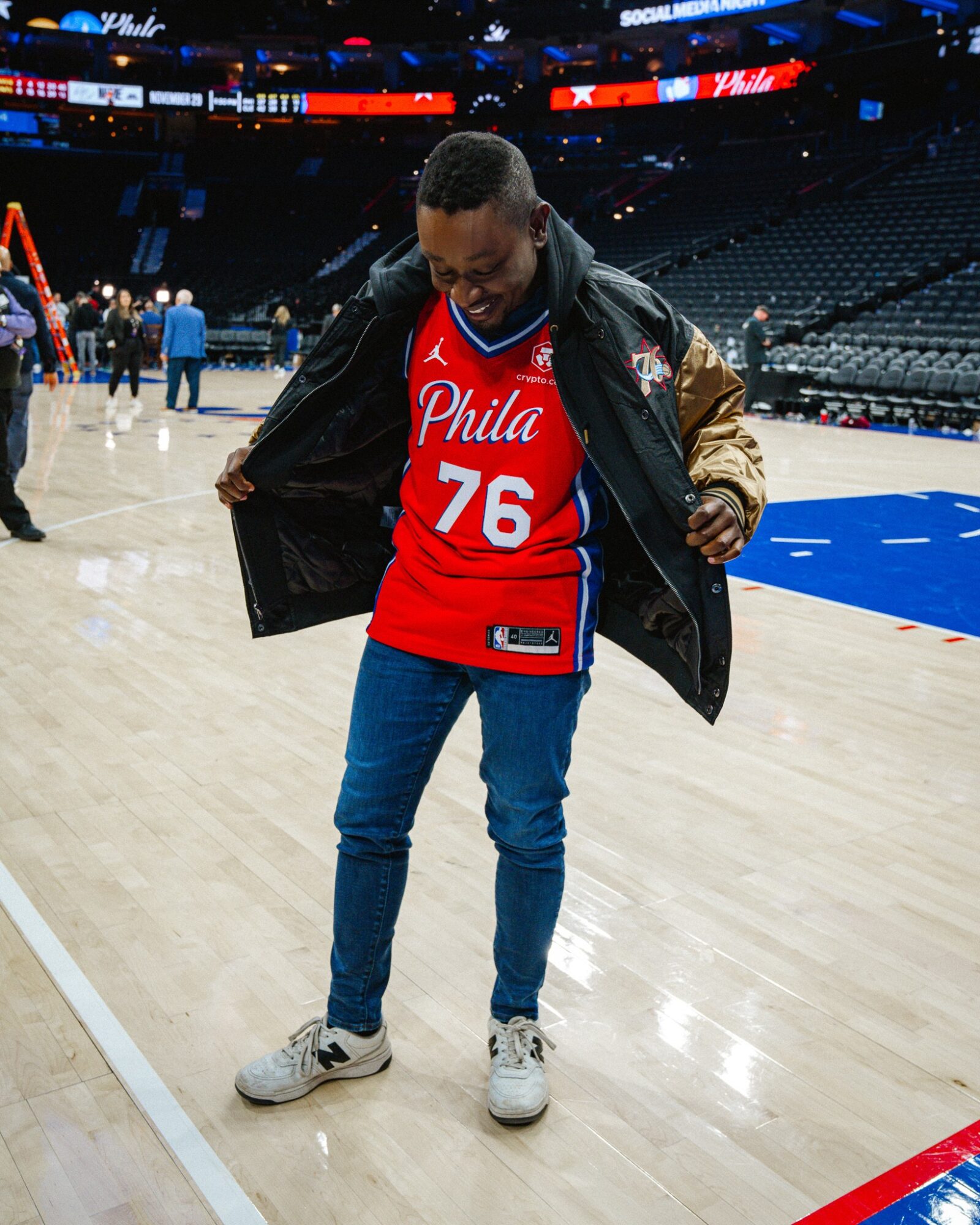 Young person standing on basketball court, wearing red Philadelphia 76ers jersey, jeans, and sneakers, adjusting jacket.