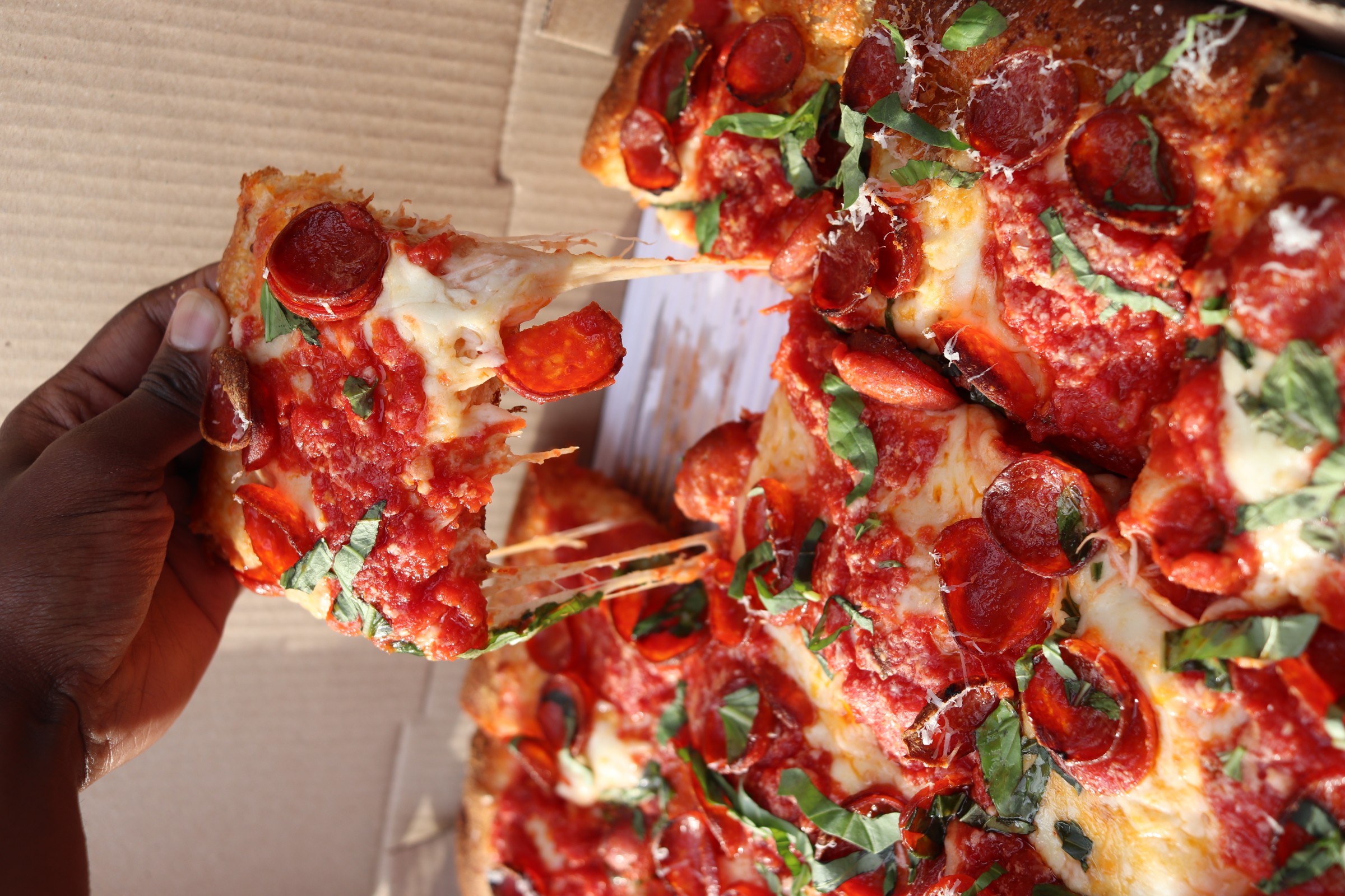 Person holding a slice of pepperoni pizza with cheese, tomato sauce, and basil, with more pizza slices in the background.