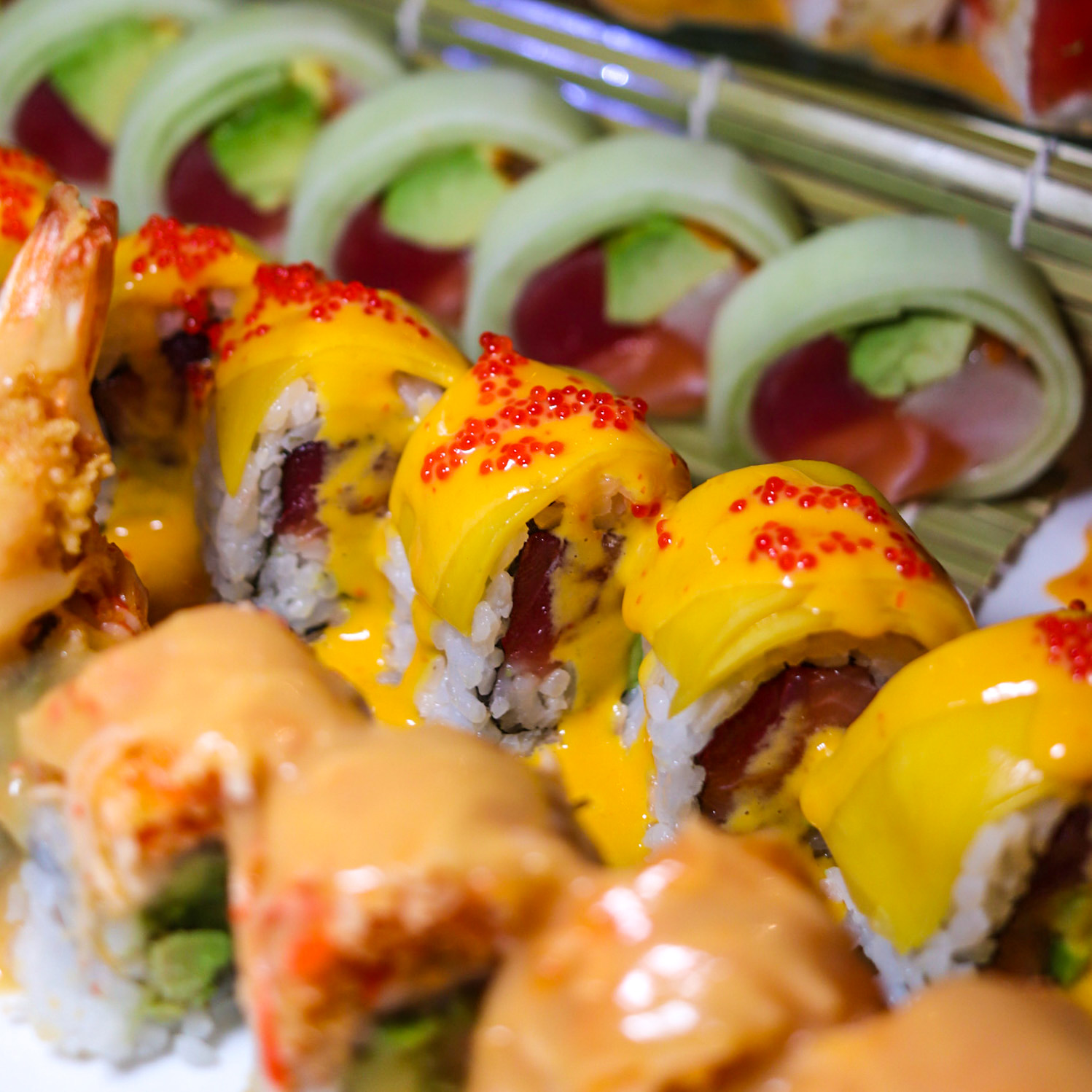 Close-up of colorful sushi rolls with yellow sauce and red fish roe, with green and pink garnishes in the background.
