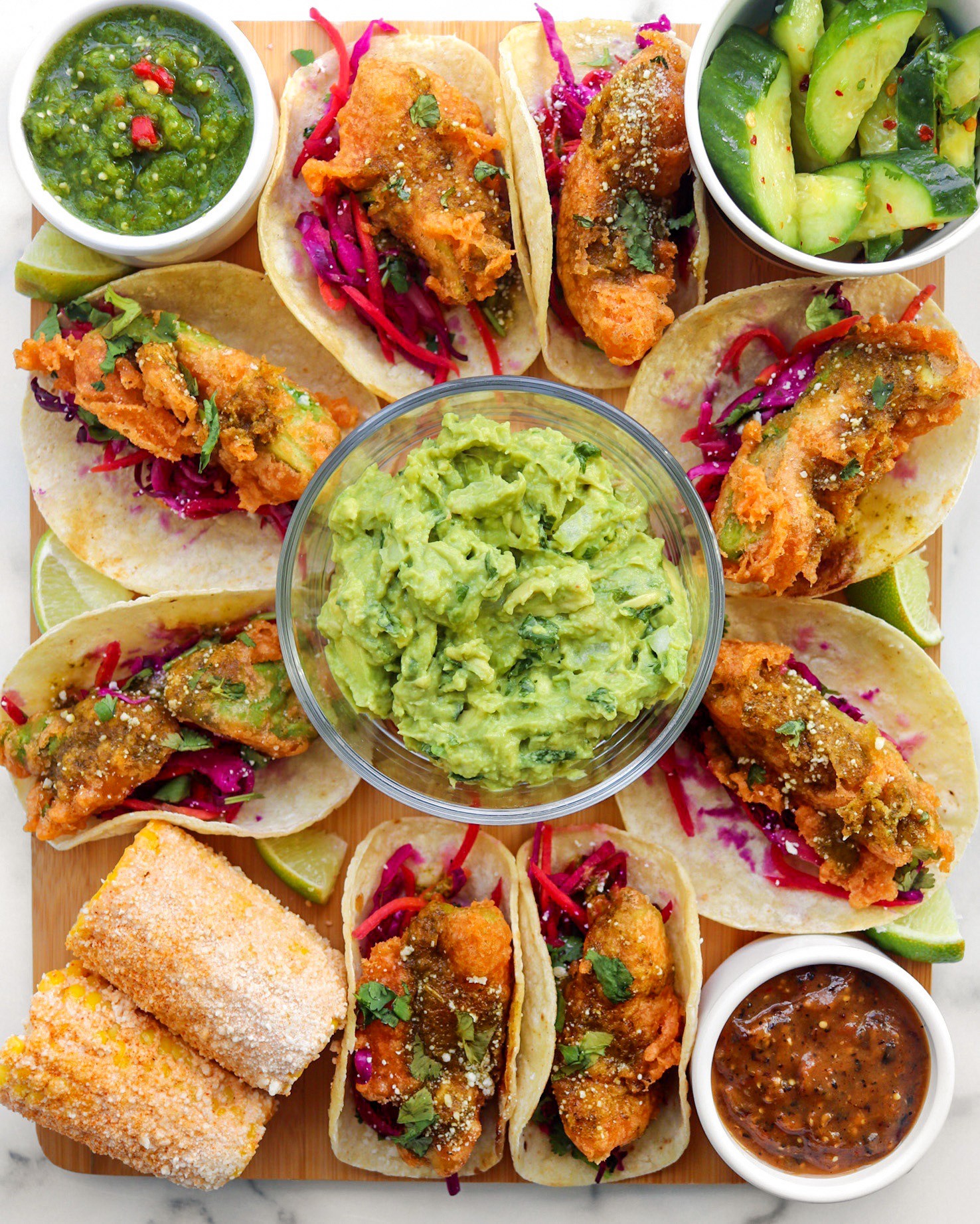 Assorted tacos with guacamole, cucumber salad, fried items, and salsa, arranged on a tray with small bowls of sauces.