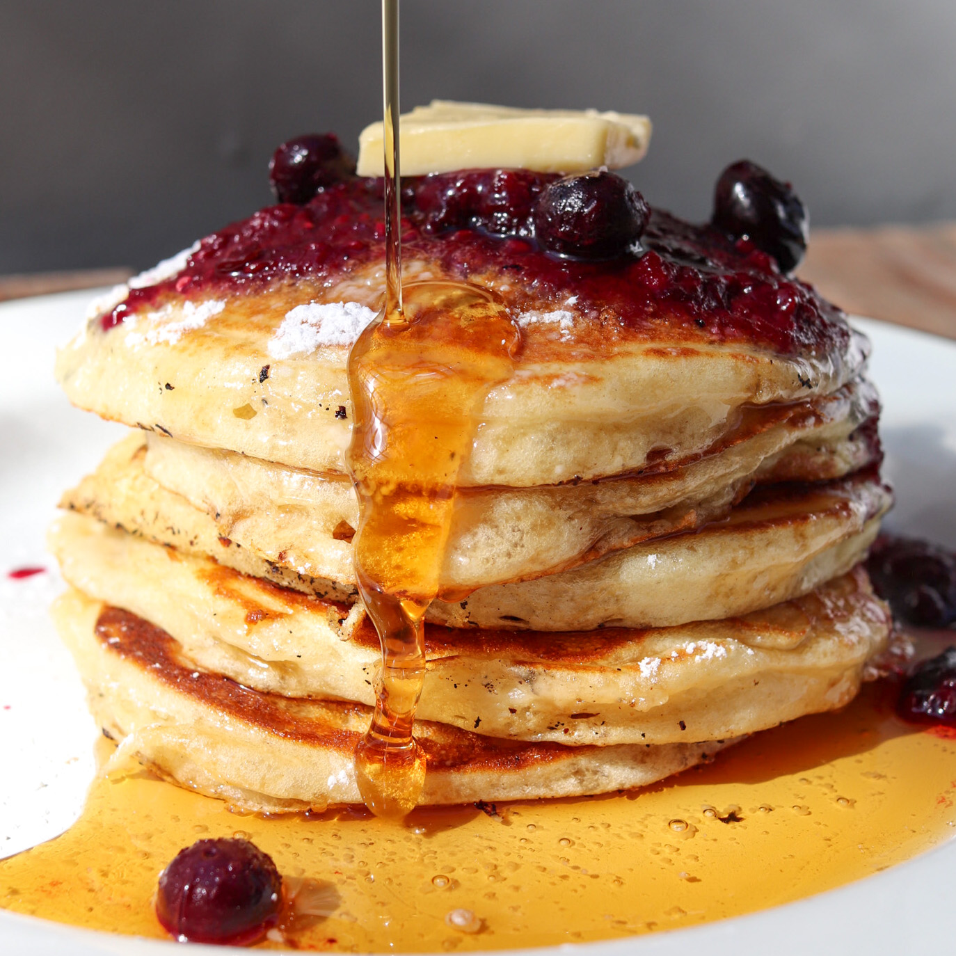Stack of four pancakes topped with berries, syrup, and a pat of butter, with syrup dripping down the sides.