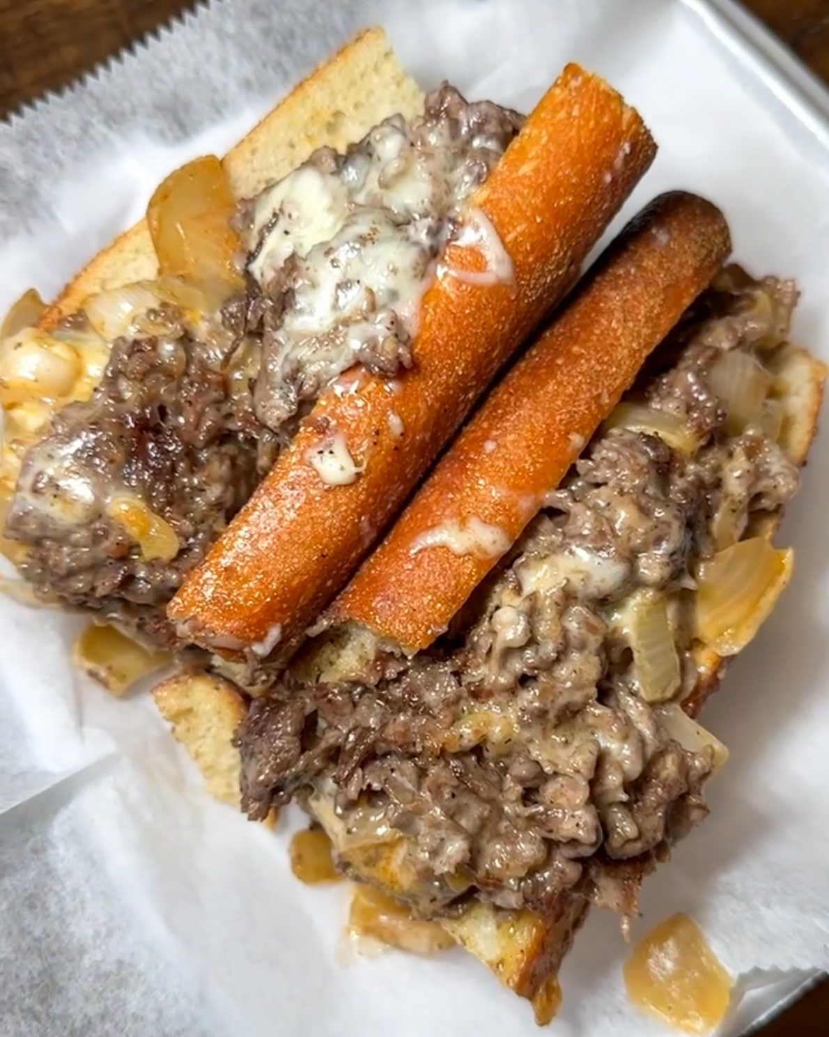 Two beef sandwiches with melted cheese, onions, and bread, served on a white paper-lined tray.