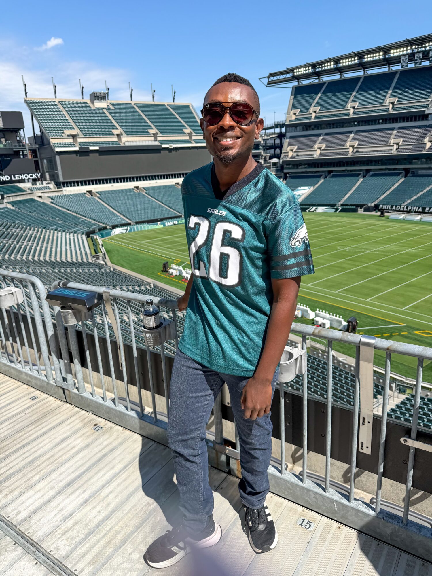 Man wearing sunglasses and a teal sports jersey with number 26, standing in a stadium with empty seats and a field behind him.
