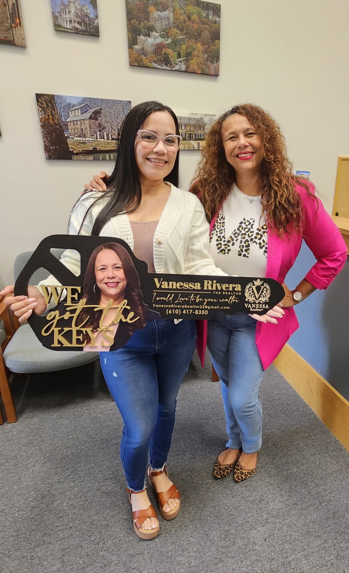 Two women standing indoors, smiling, holding a sign, with framed pictures on the wall behind them.