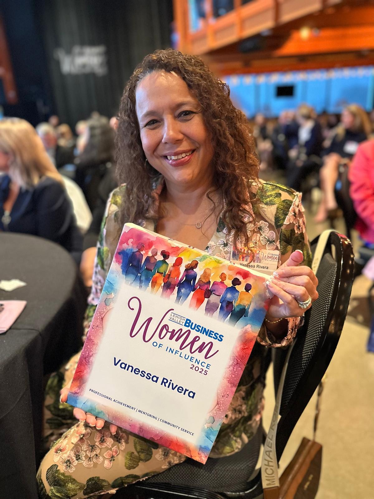 Woman smiling holding a certificate at an event, with a crowd in the background.