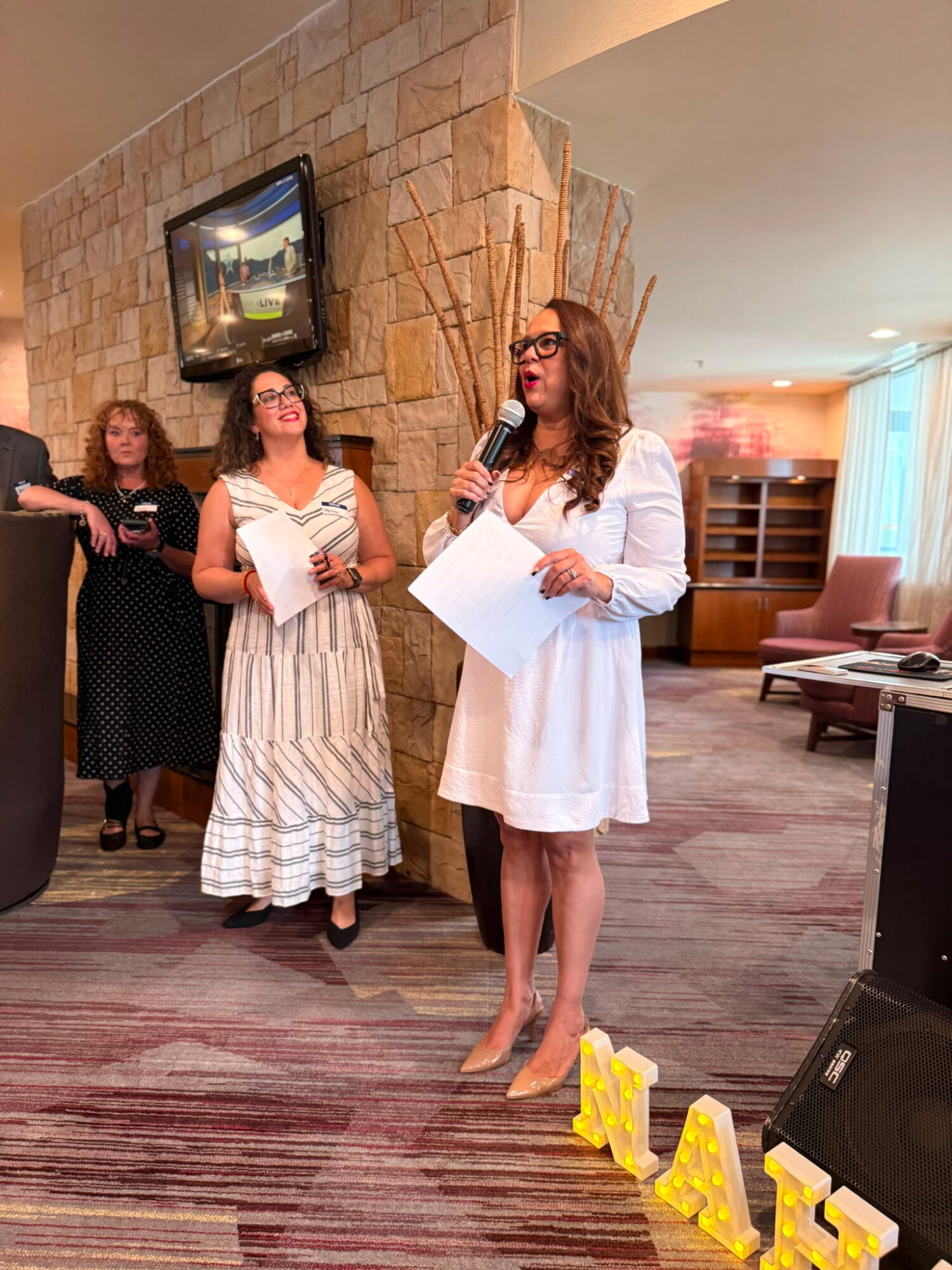 Woman in white dress speaking into microphone, holding papers, with three women in background, in a room with a TV and seating area.