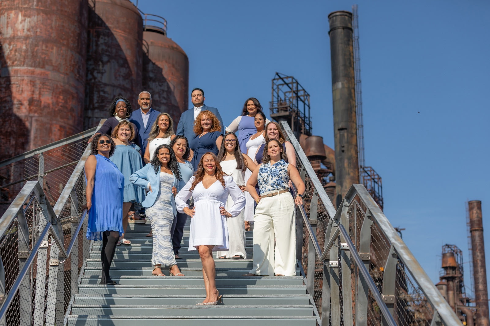 Group of women standing on industrial stairs outdoors, with factory structures and blue sky in background.