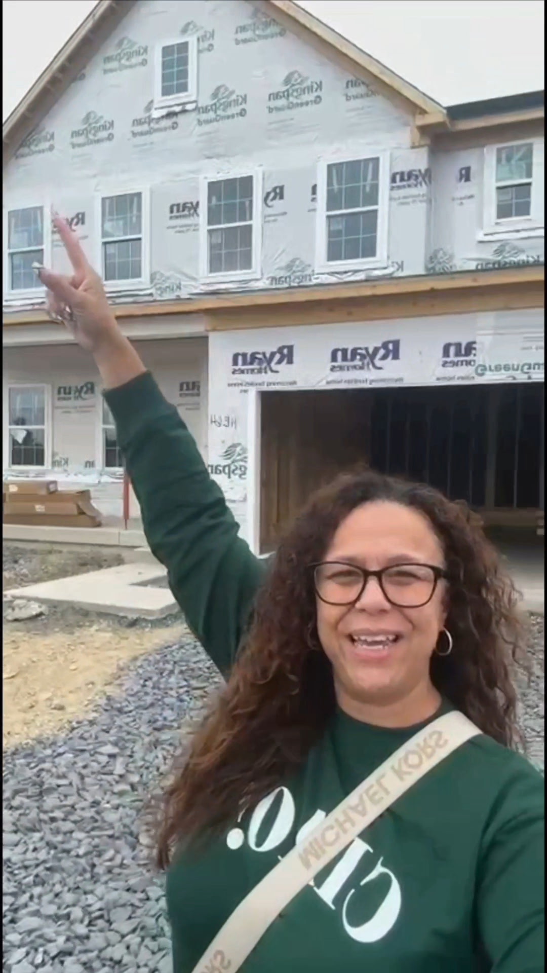 Woman with glasses smiling and making a peace sign in front of a house under construction.