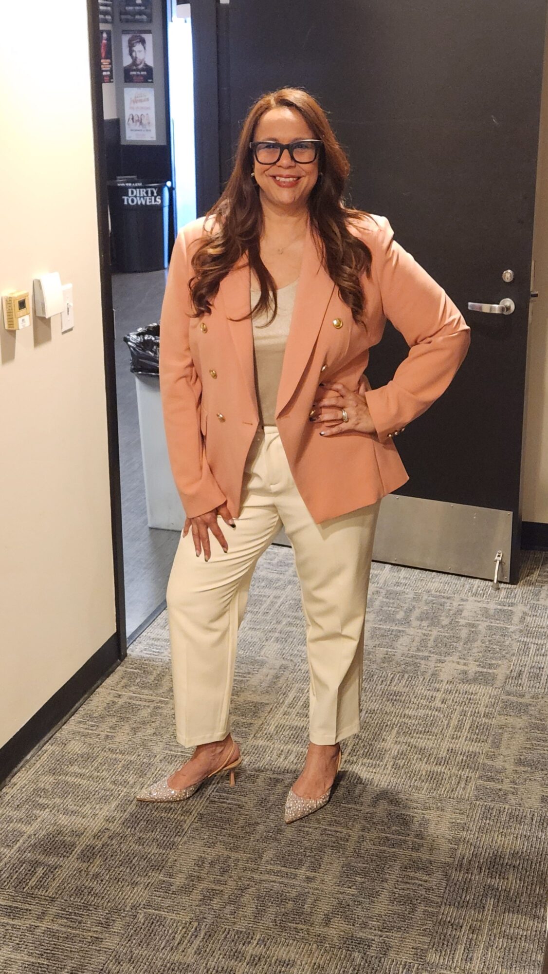 Woman with long hair and glasses standing in hallway, smiling, wearing a peach blazer, cream top, and beige pants.