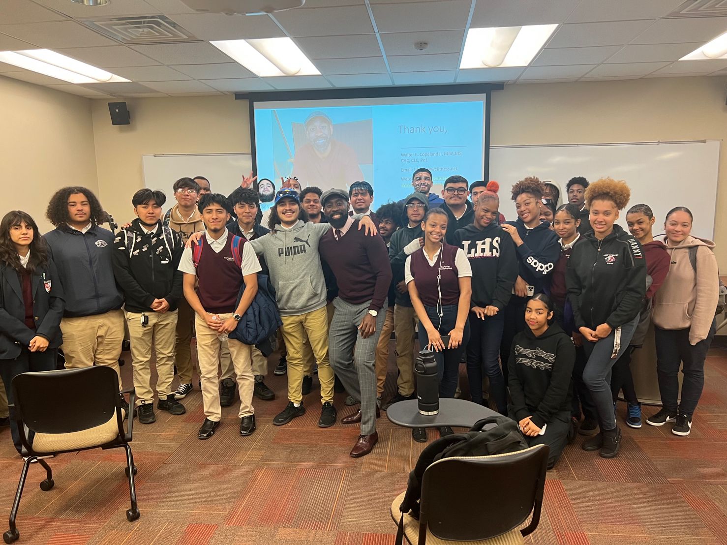 Group of students and teachers in a classroom, standing in front of a large screen, smiling for a photo.