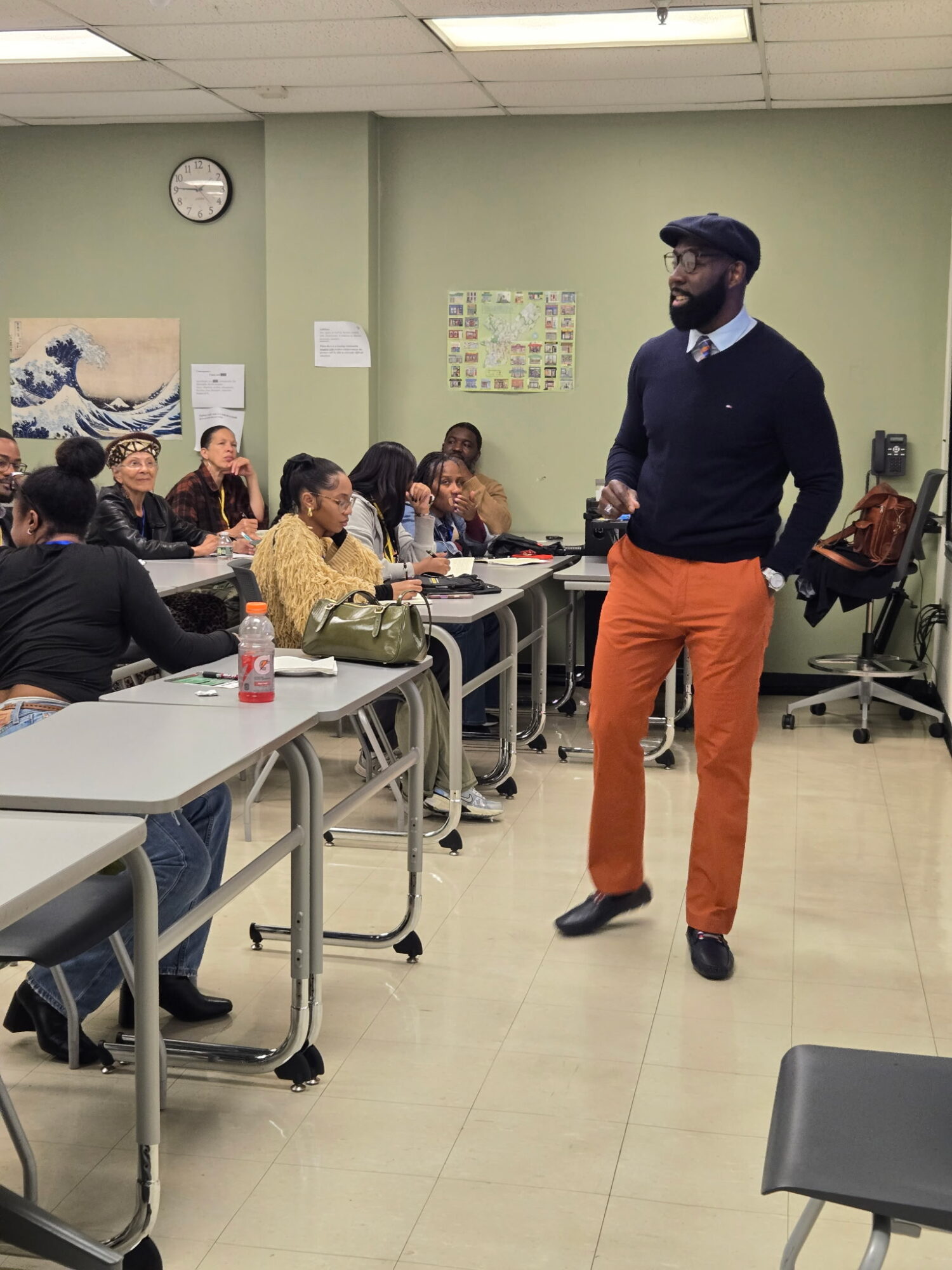 Man standing in front of seated group in classroom, wearing dark sweater, orange pants, and cap, with a whiteboard behind him.