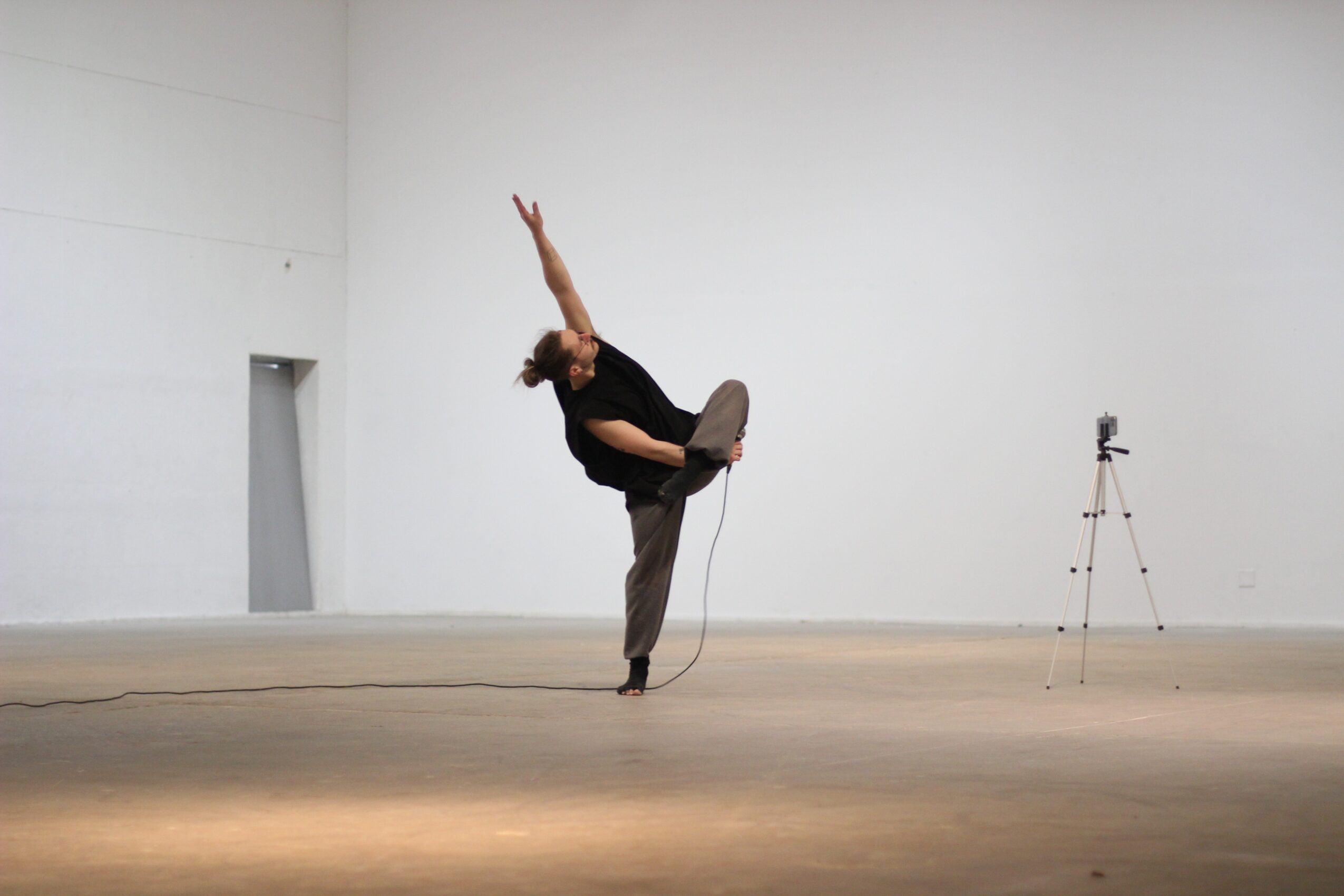 Person balancing on one leg with arms raised in a large, empty room, with a tripod in the background.