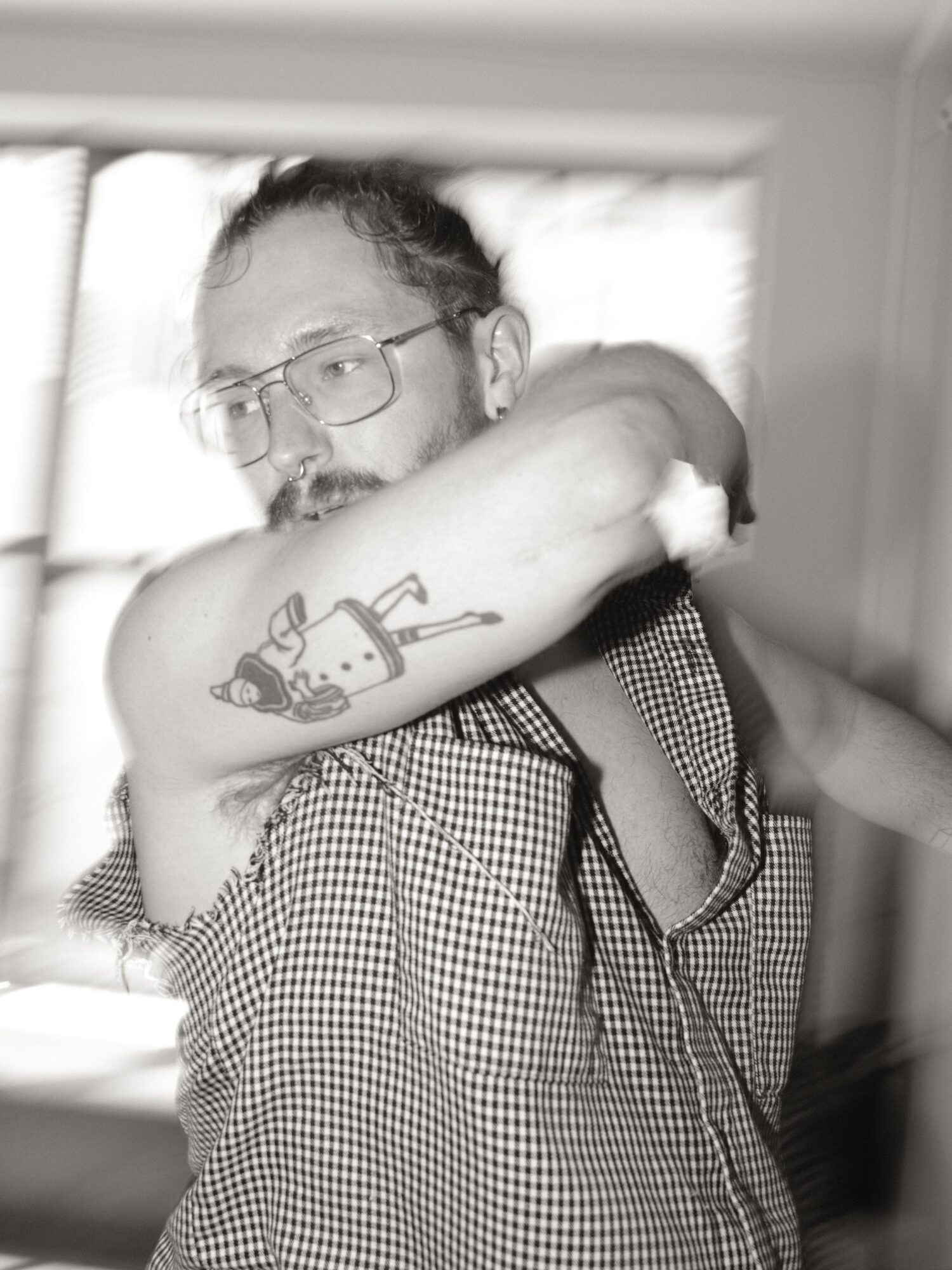 Person with glasses and a tattoo on arm, lying on side, in a checkered shirt, in a room with window blinds.