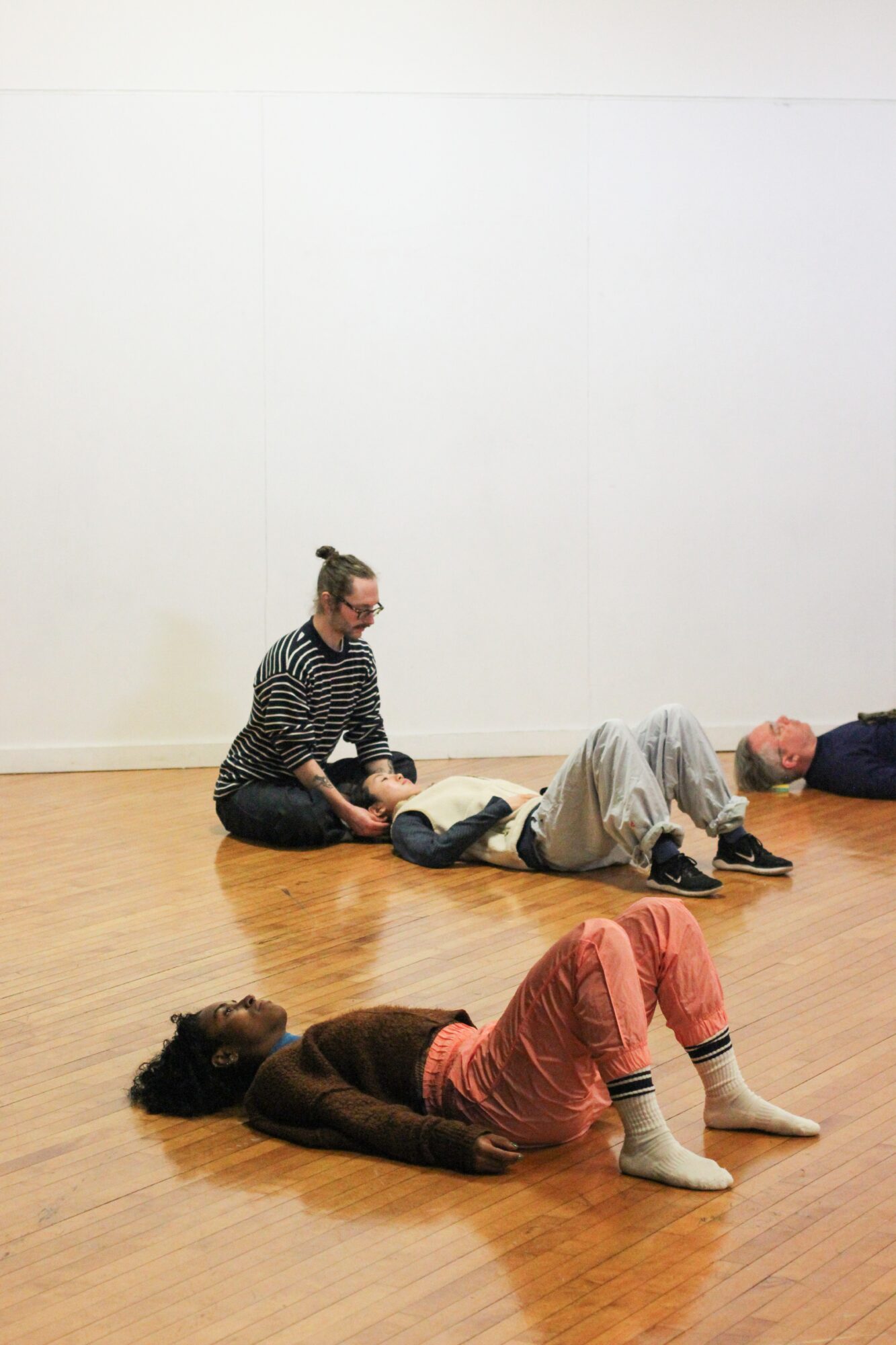 Four people lying on the floor in a room with a white wall and wooden floor, one person sitting nearby.