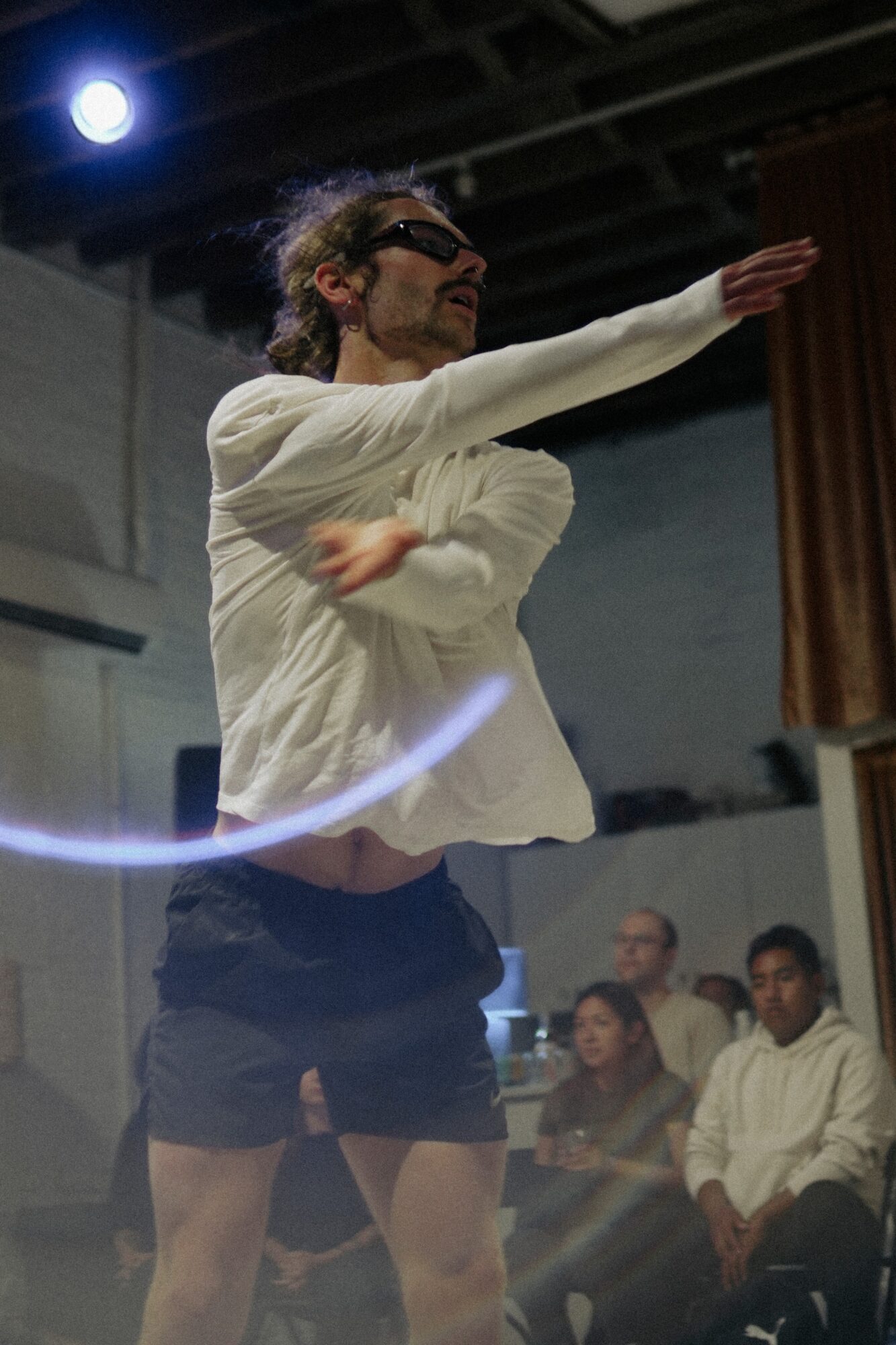 Person with curly hair and glasses dancing indoors, wearing a white long-sleeve shirt and black shorts, with audience in background.