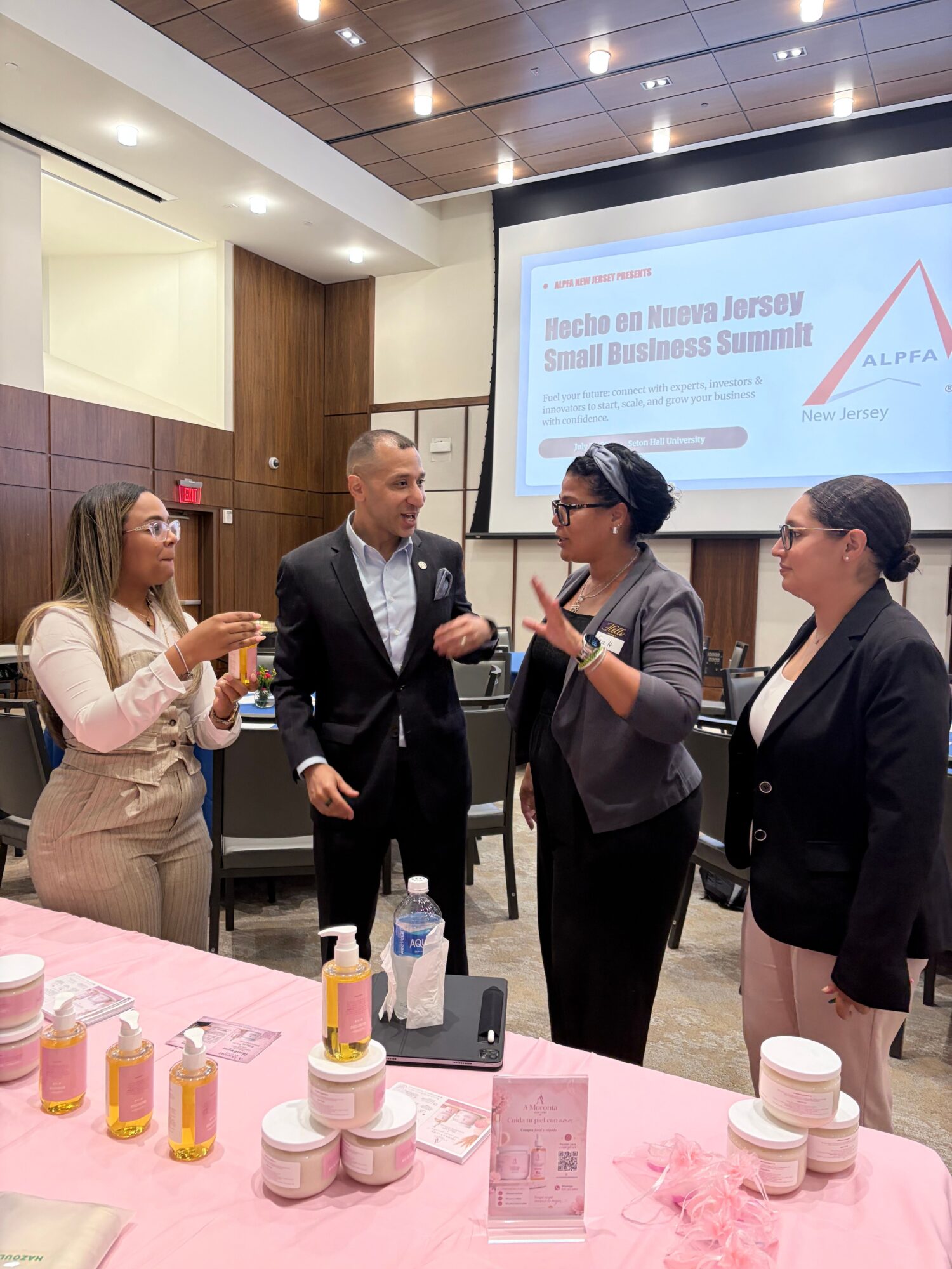 Three people standing and talking in a conference room with a presentation screen behind them, a pink table with drinks and snacks in front.