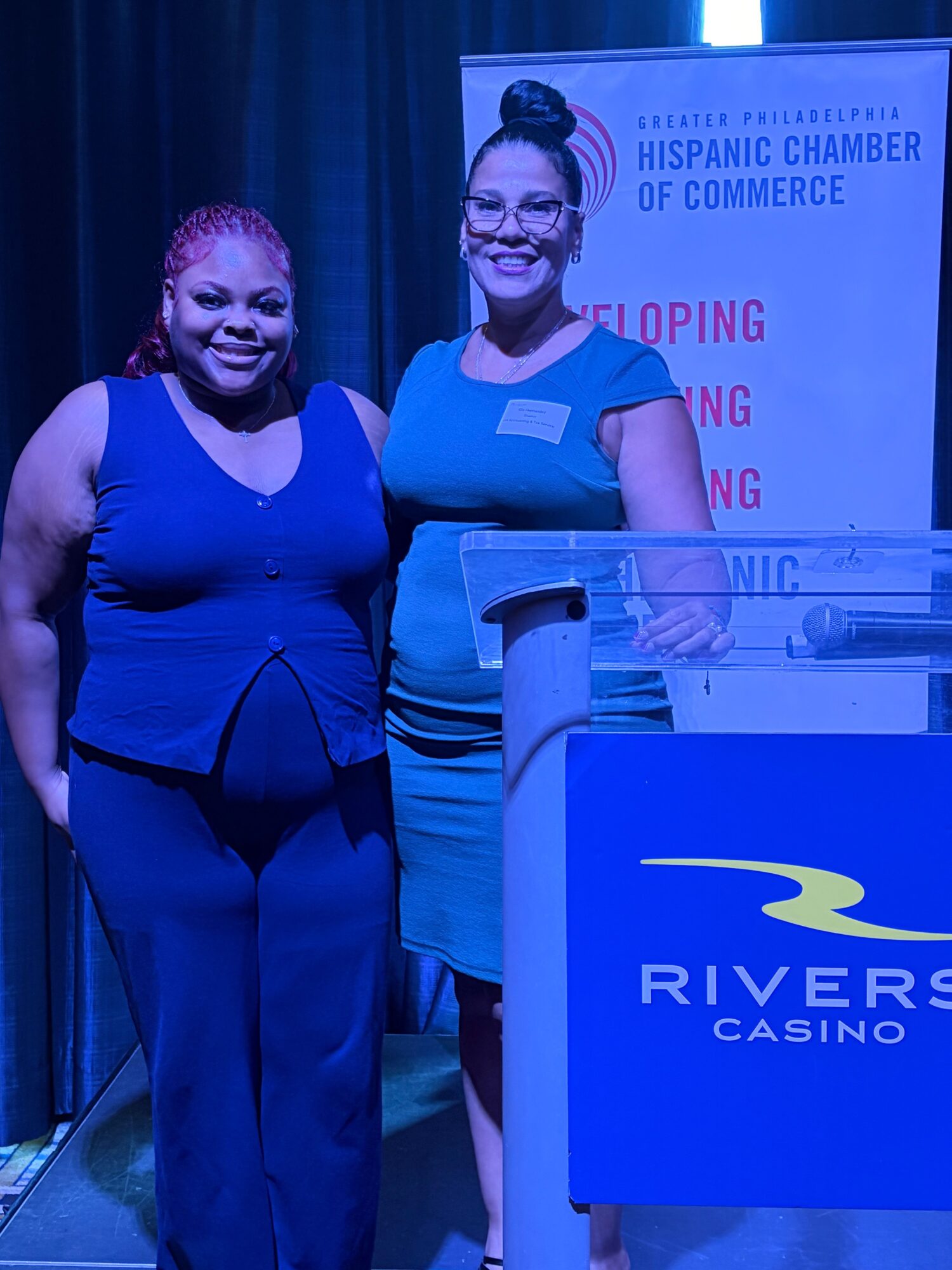 Two women standing behind a podium at a conference, smiling, with a blue and yellow backdrop and a sign reading 'Rivers Casino'.