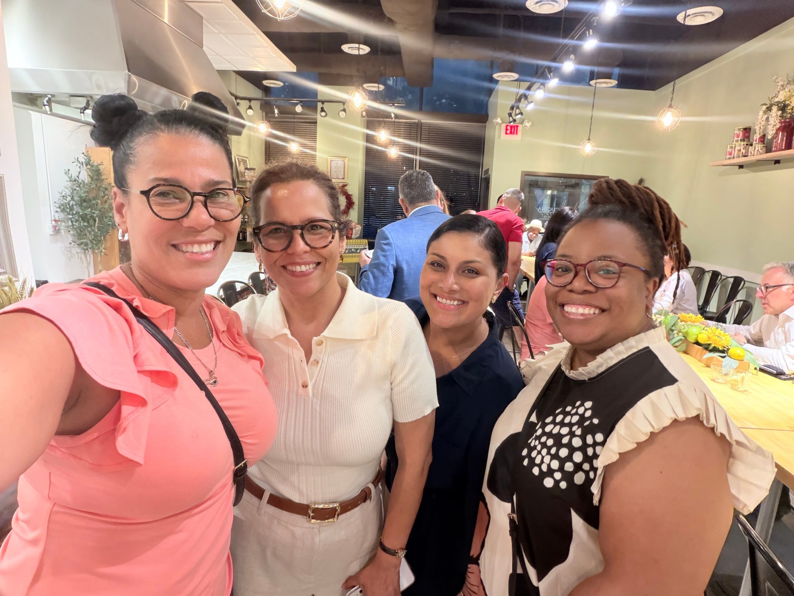 Four women smiling and posing together in a restaurant or event space, with tables and decorations in the background.