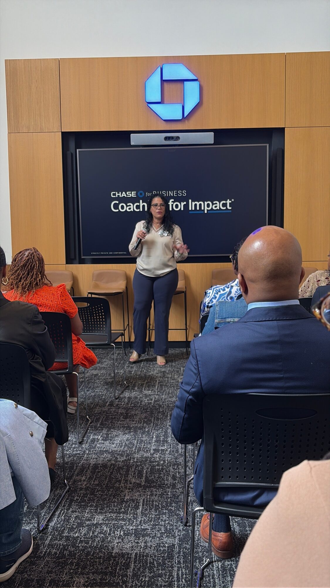 Woman speaking to an audience in a conference room with a screen displaying 'Coach for Impact' and a Chase logo above.