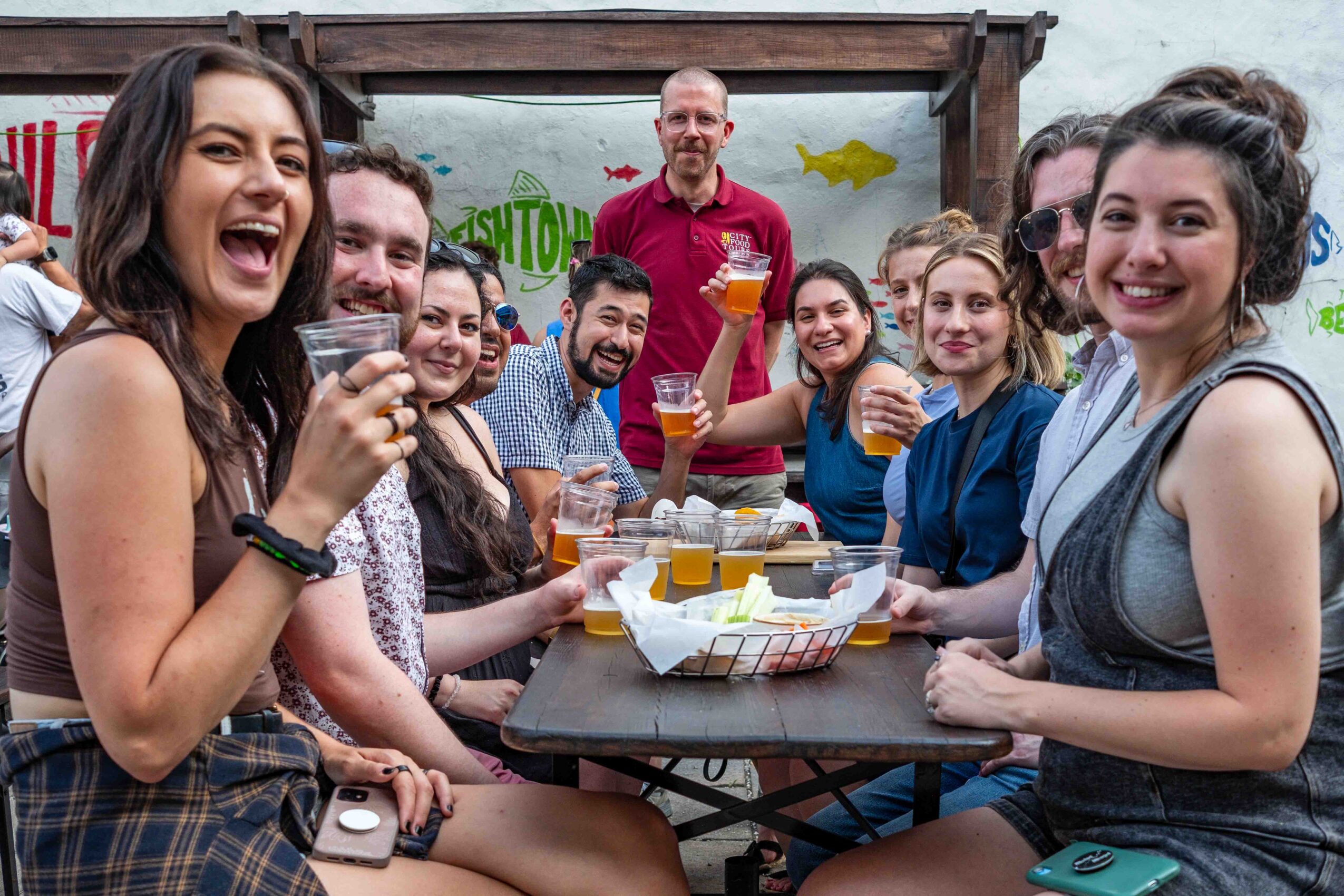 Group of people sitting at a table outdoors, smiling and holding drinks, with a man standing behind them. 20 words.
