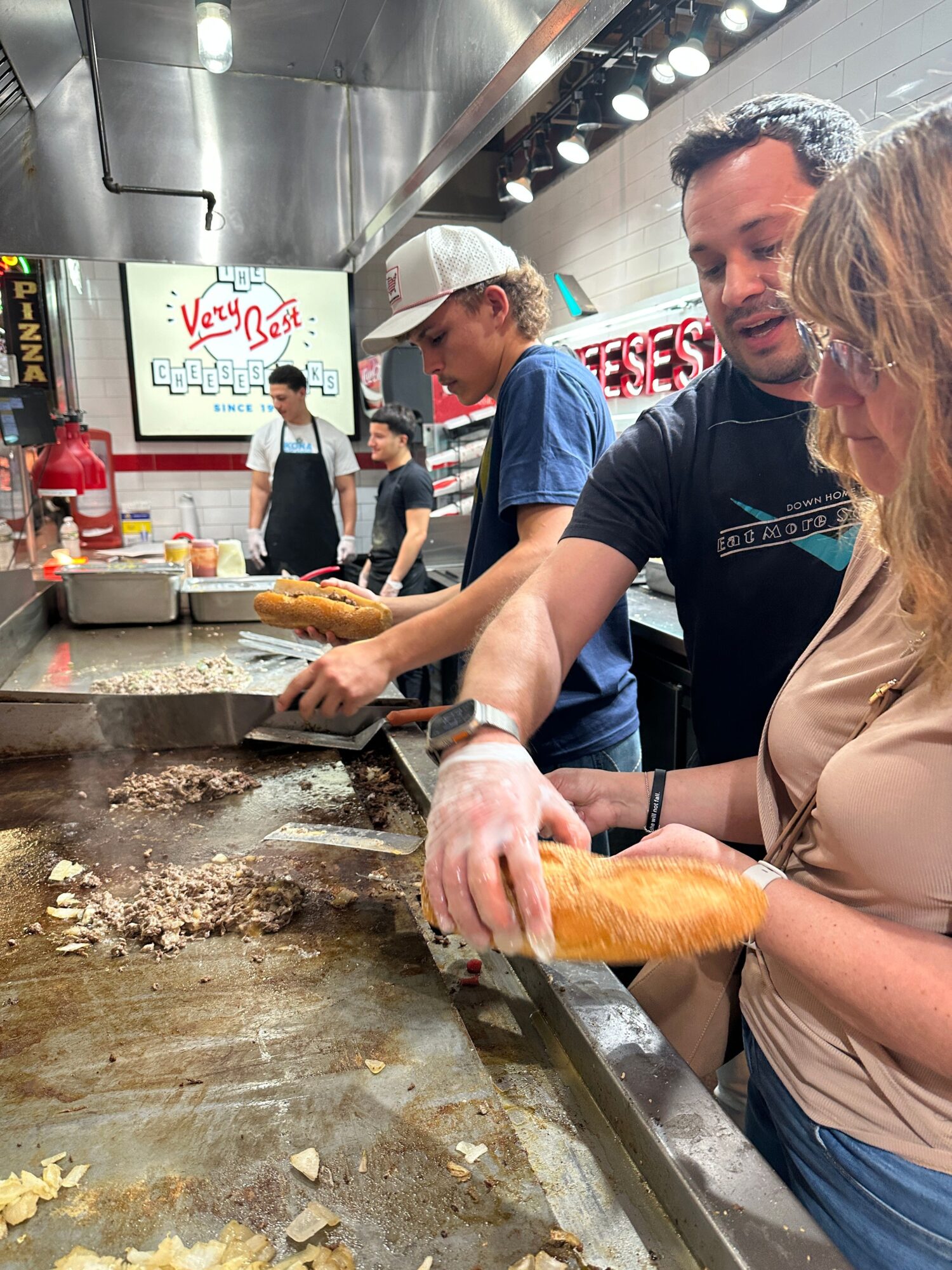 Two people preparing food at a counter, with a man in a black shirt and a woman in a beige top, in a busy kitchen or food stall.