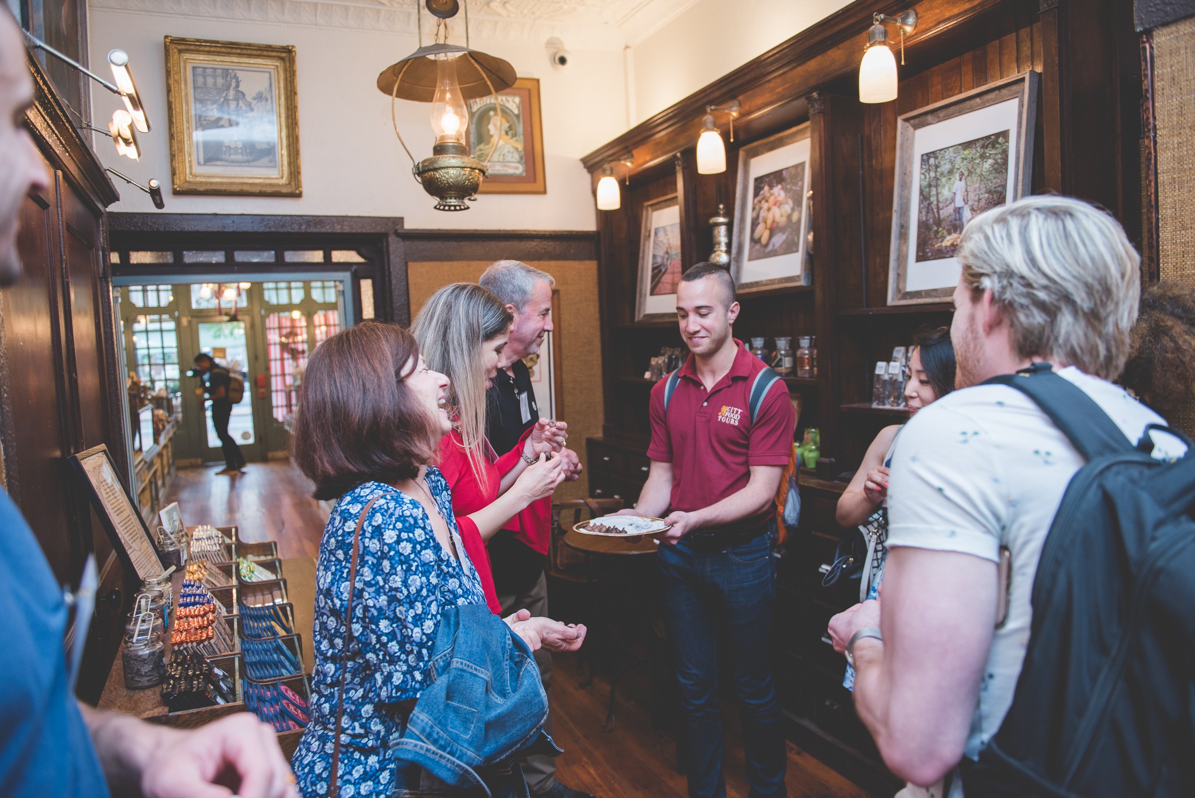 Group of people inside a room with wooden walls, framed pictures, and a person behind a counter talking to others.