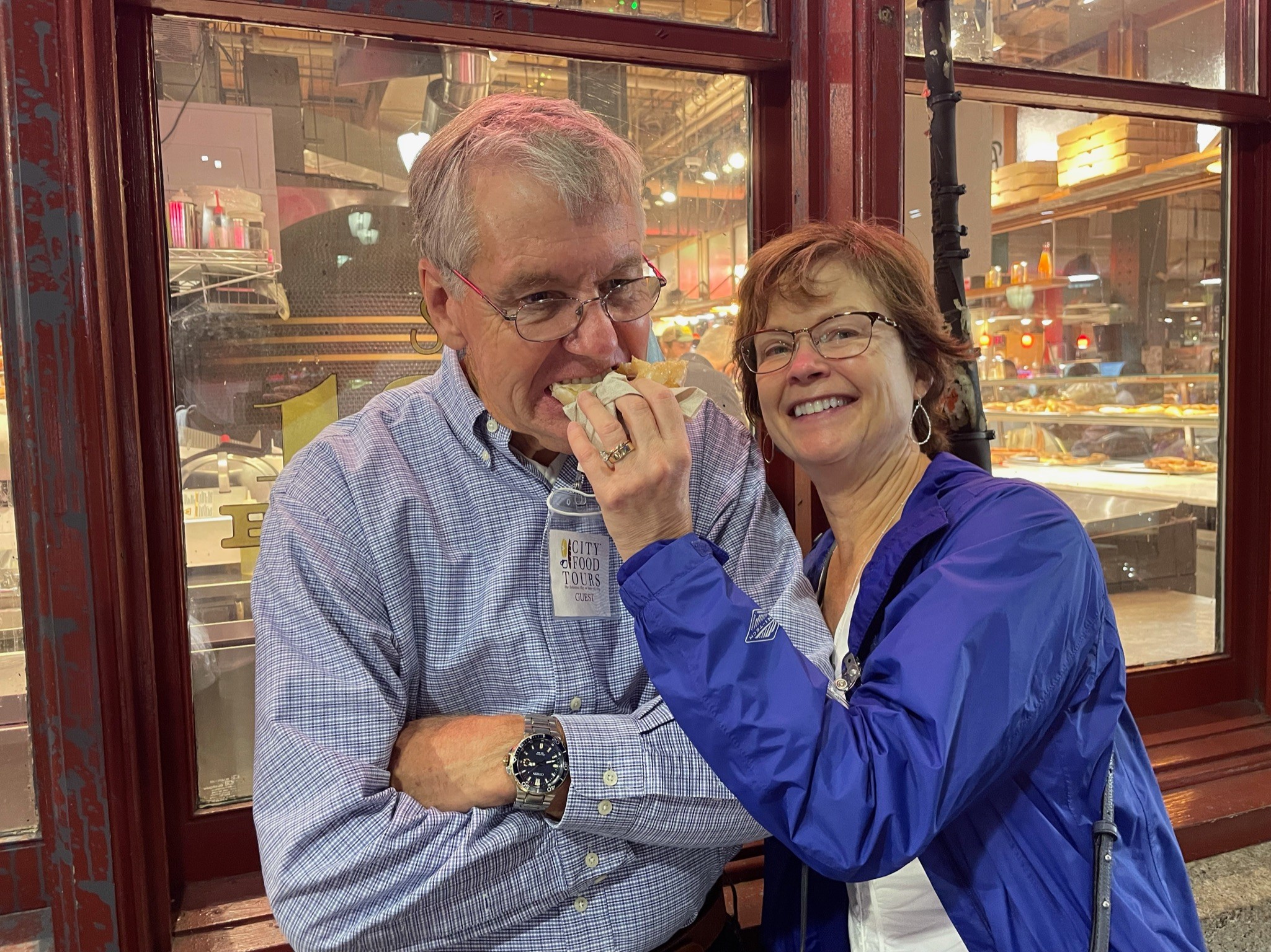 Two smiling people, one feeding the other, inside a restaurant with large windows and city lights outside.