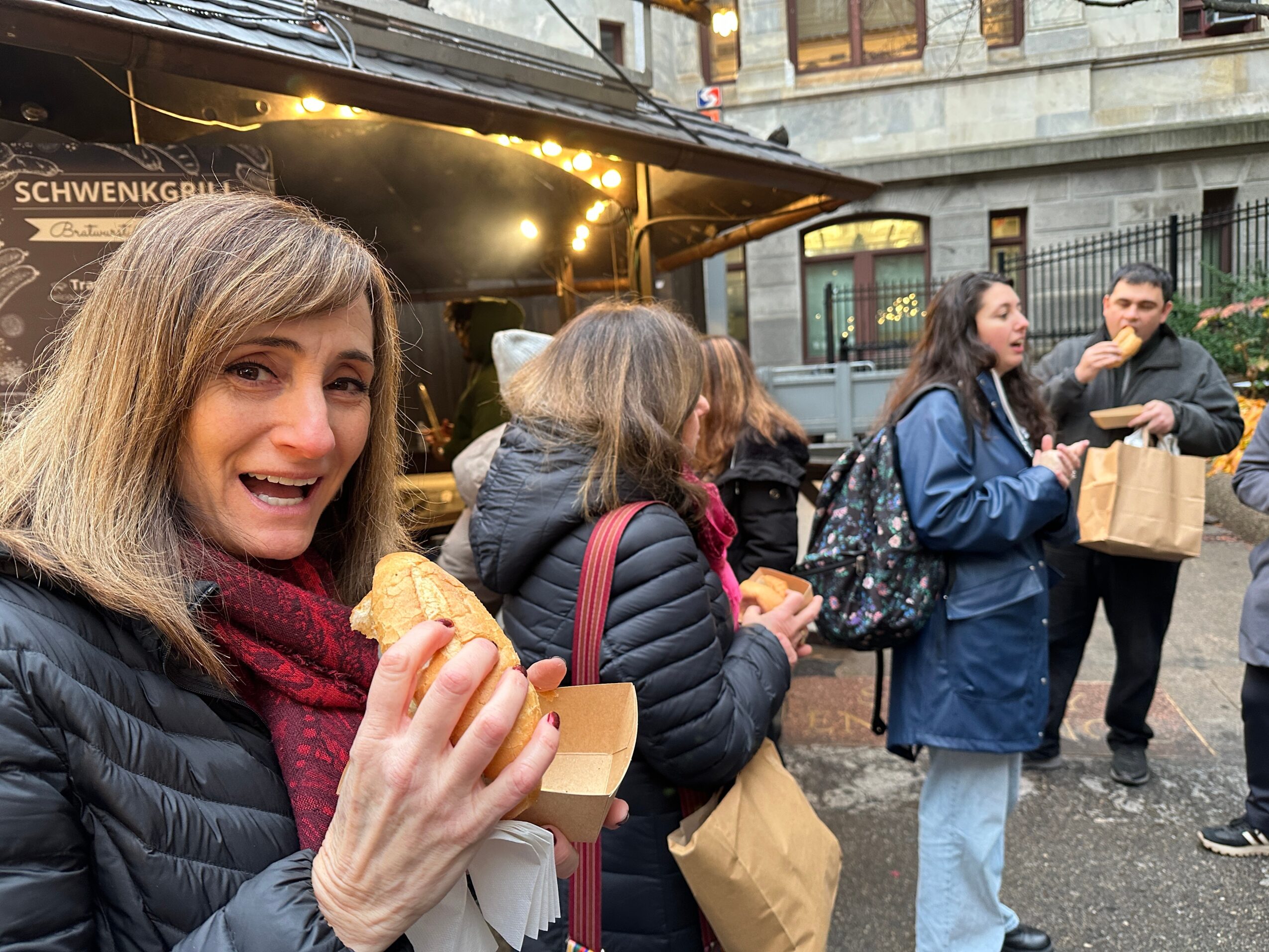 Woman smiling and holding a pastry in front of a food stand outdoors, with people in the background.