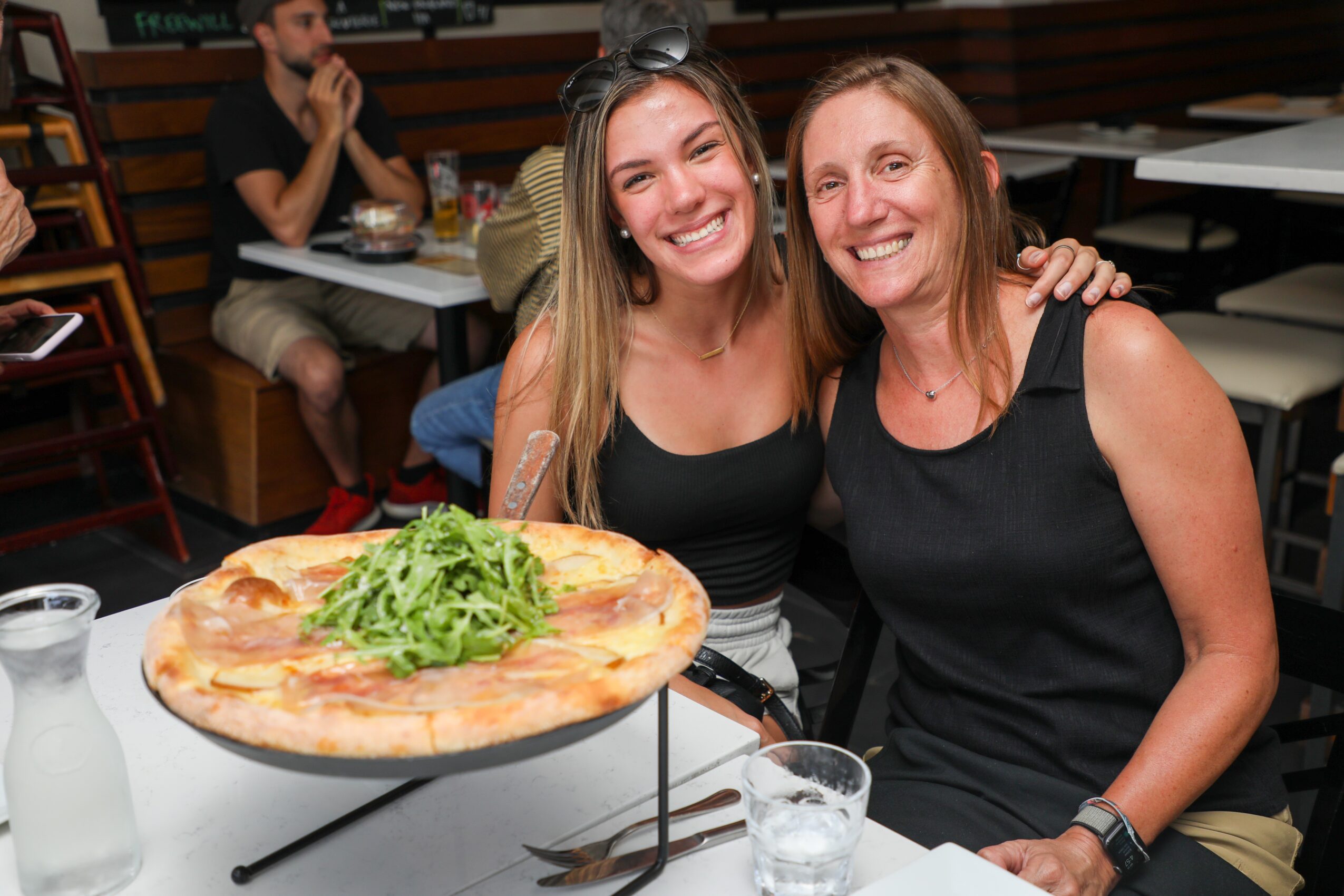 Two women smiling and sitting at a table with a pizza and drinks, in a restaurant setting.
