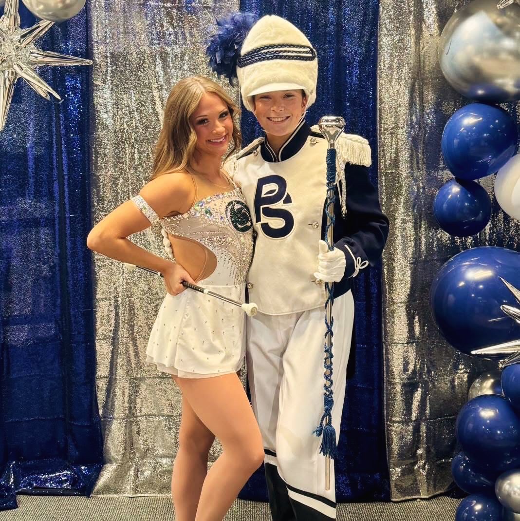 A girl in a white dress and a boy in a marching band uniform stand together, smiling, with festive decorations behind them.