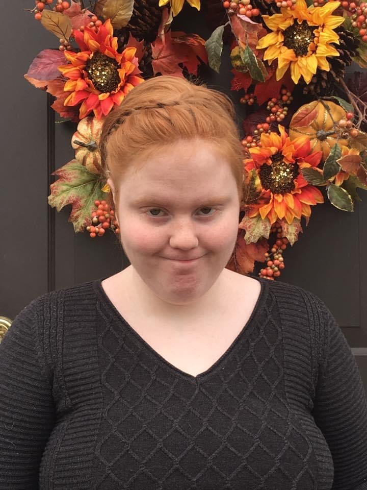 Young woman with red hair in a bun, smiling, wearing a black sweater, standing in front of autumn-themed floral display.