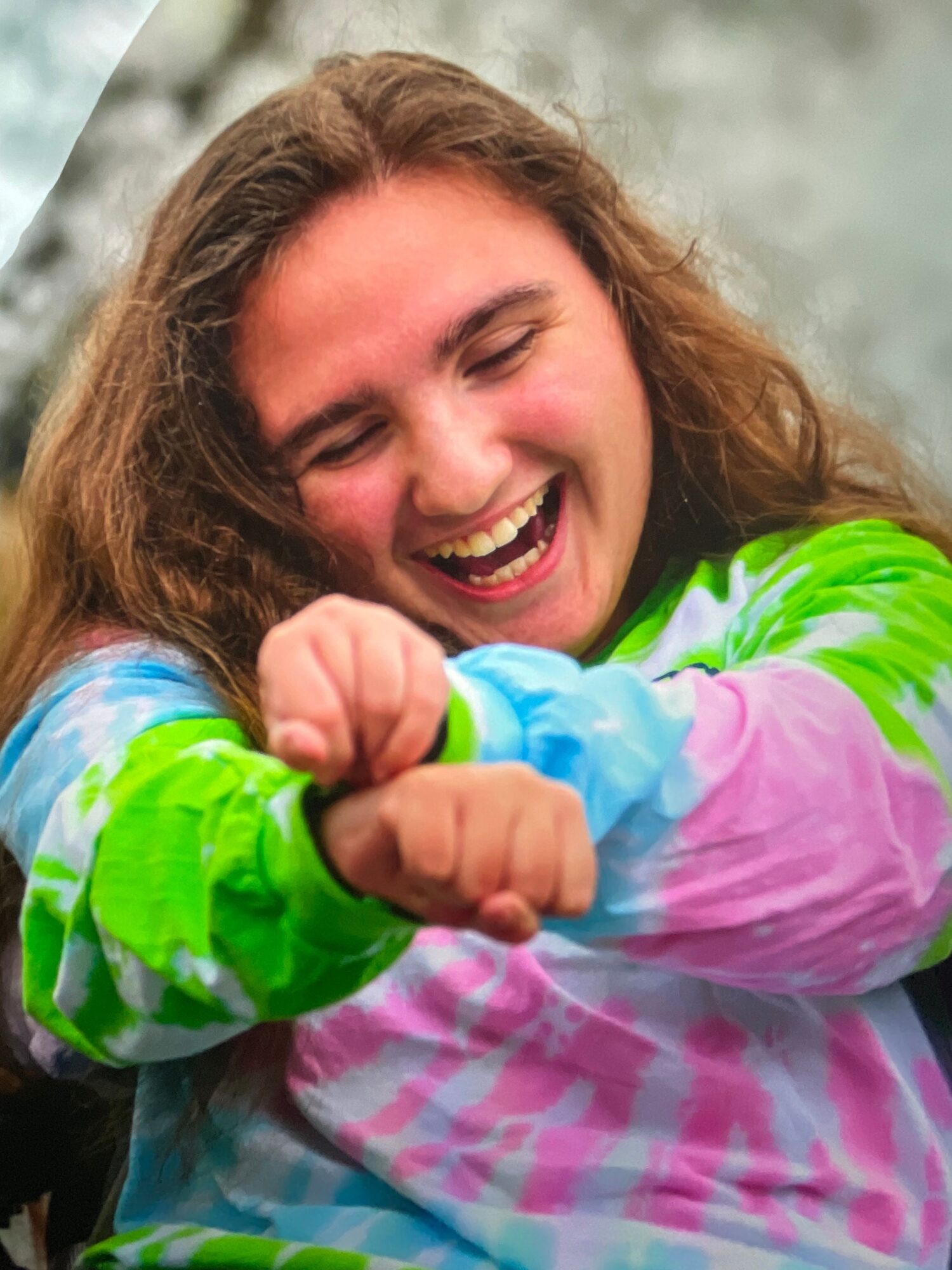 Person laughing outdoors, wearing colorful tie-dye clothing, with hands clenched in excitement.