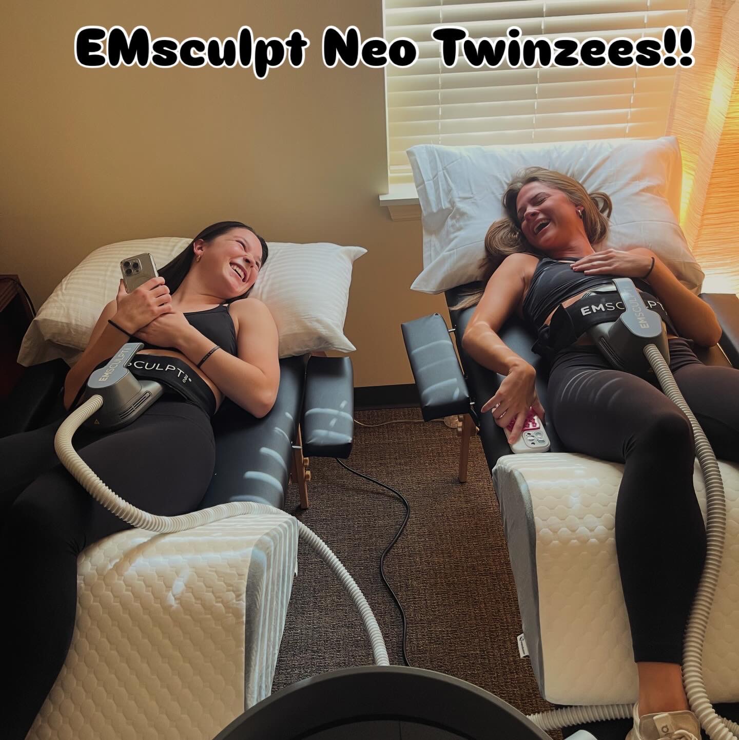 Two women lying on medical chairs, smiling, with equipment attached, in a room with a window and blinds.