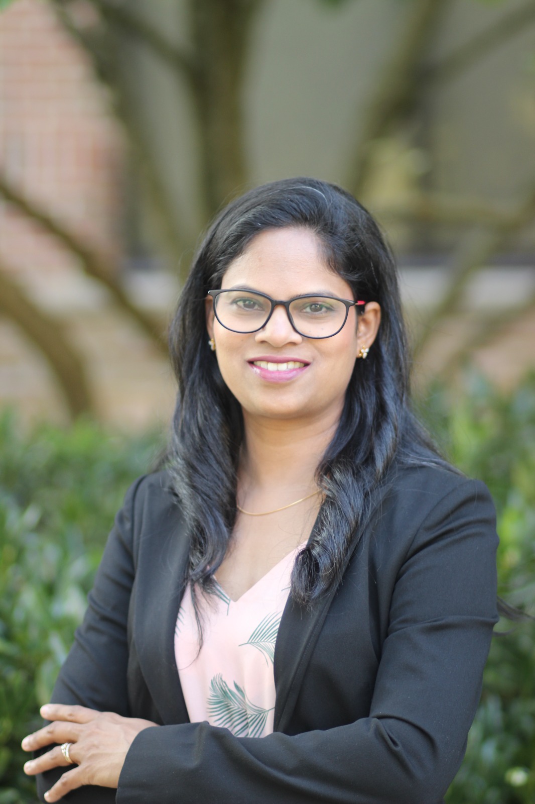 Woman with glasses and black hair smiling outdoors, wearing a black blazer and light-colored blouse, with trees and greenery in background.