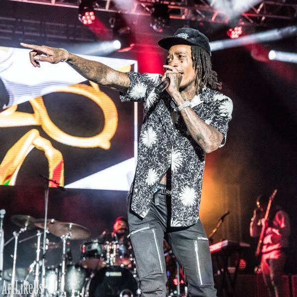 Performer singing into microphone on stage with band, pointing to the audience, colorful lights in background.