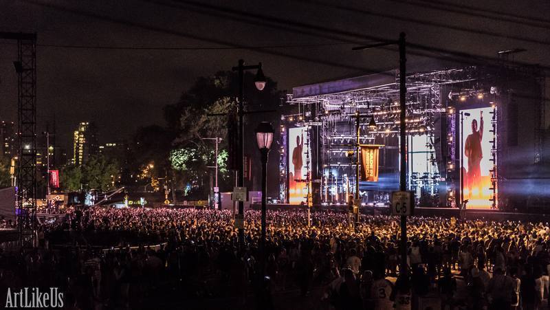 Large outdoor concert stage illuminated at night with a crowd in front, city lights in the background.