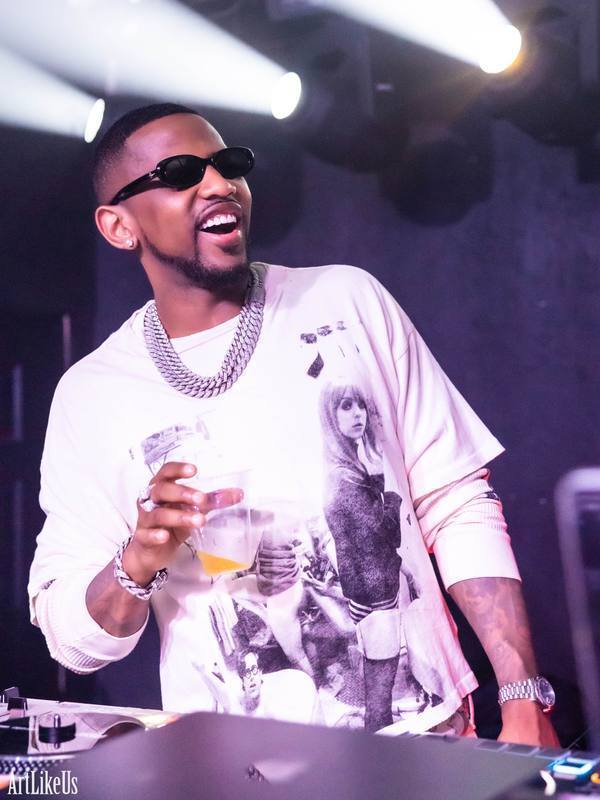 Man wearing sunglasses and a graphic t-shirt, holding a drink, smiling at a party or event, with stage lights overhead.