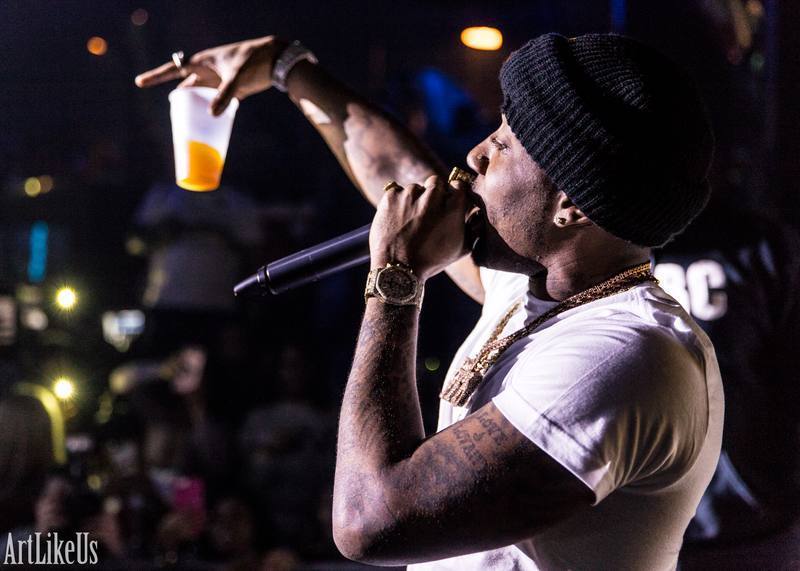 Performer with a beanie and tattoos sings into a microphone, holding a drink in a dark, lively setting.