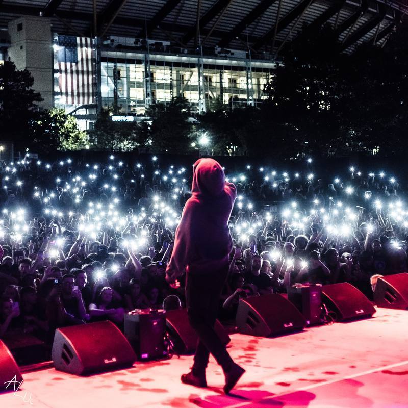 Performer on stage facing a large crowd with lights, in front of a modern building at night.