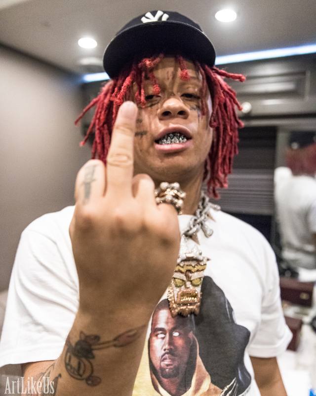 Person with red dreadlocks, wearing a black cap, showing middle finger, in an indoor setting, with a graphic t-shirt.