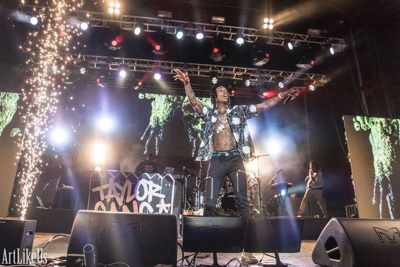 Performer on stage with arms outstretched, bright lights and digital screens behind, fireworks on the left, audience in front